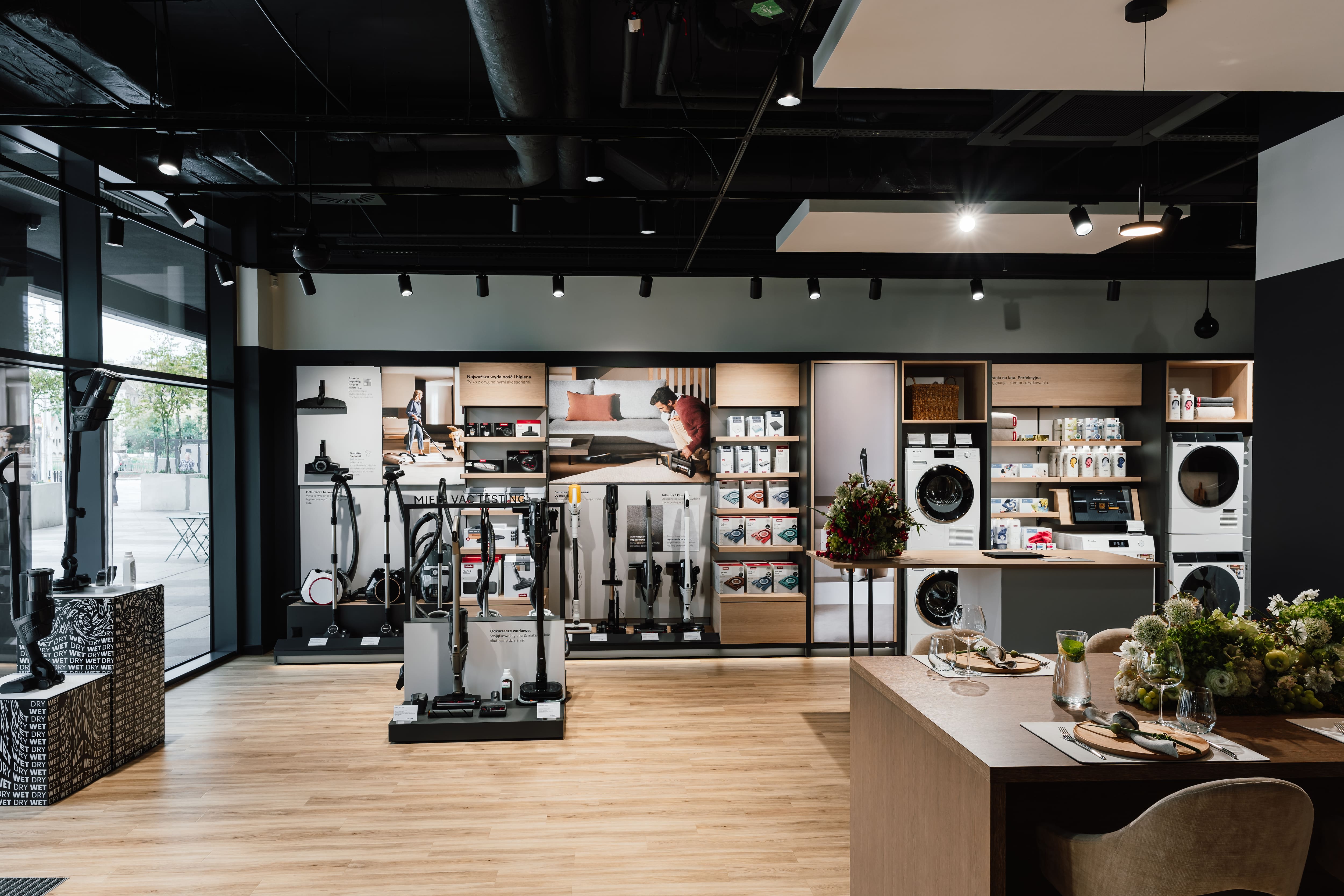 Modern appliance showroom displaying vacuums, washers, dryers, and accessories with demo island and seating.