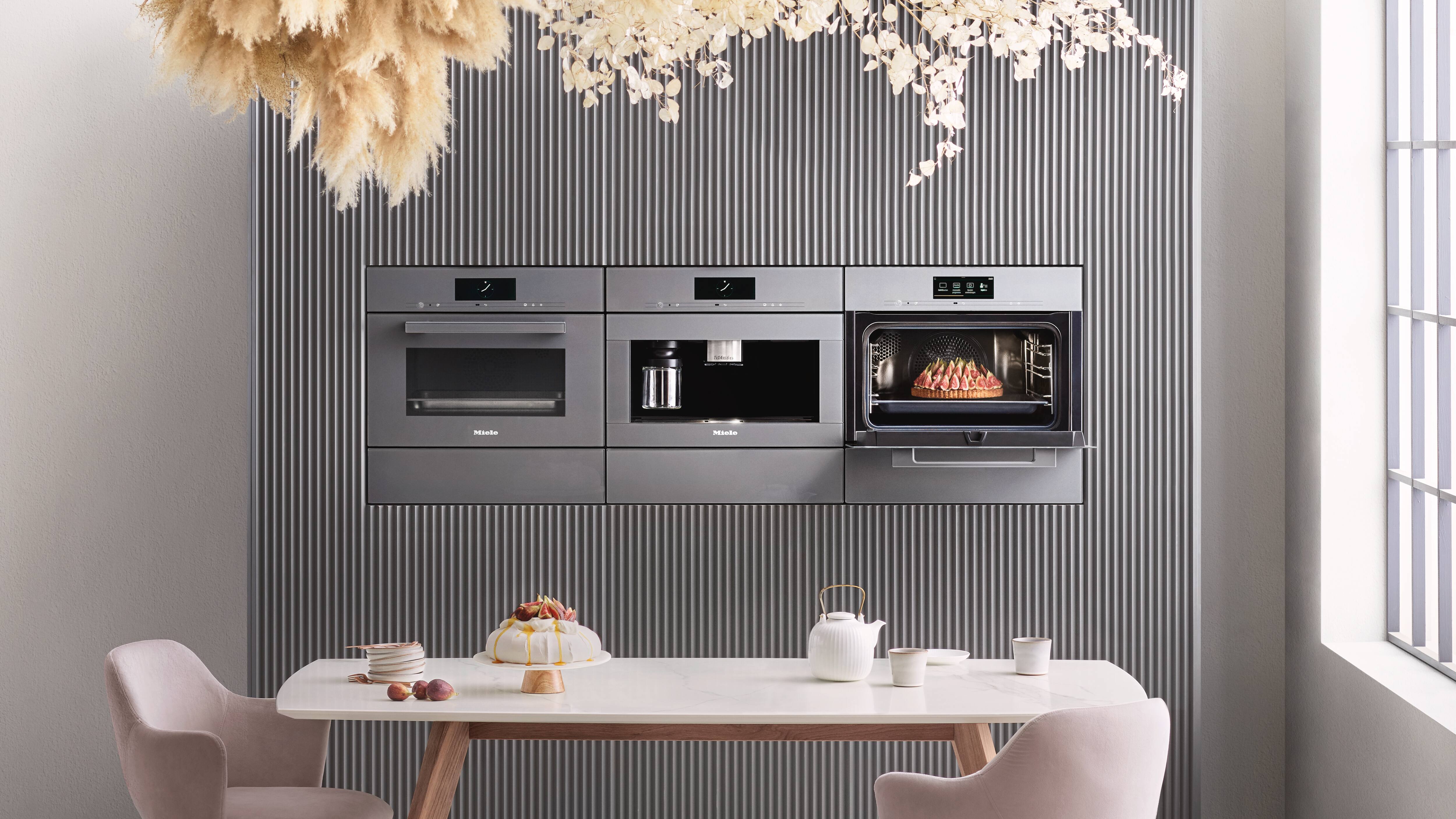 Elegant modern kitchen with three built-in stainless appliances in a ribbed gray wall—microwave/oven, built-in coffee maker, and an oven baking a roast—above a white marble dining table with wooden legs, blush chairs, a teapot, and a frosted cake; pampas and florals hang overhead with daylight from a large window on the right. Elegant modern kitchen with three built-in stainless appliances in a ribbed gray wall—microwave/oven, built-in coffee maker, and an oven baking a roast—above a white marble dining table with wooden legs, blush chairs, a teapot, and a frosted cake; pampas and florals hang overhead with daylight from a large window on the right.