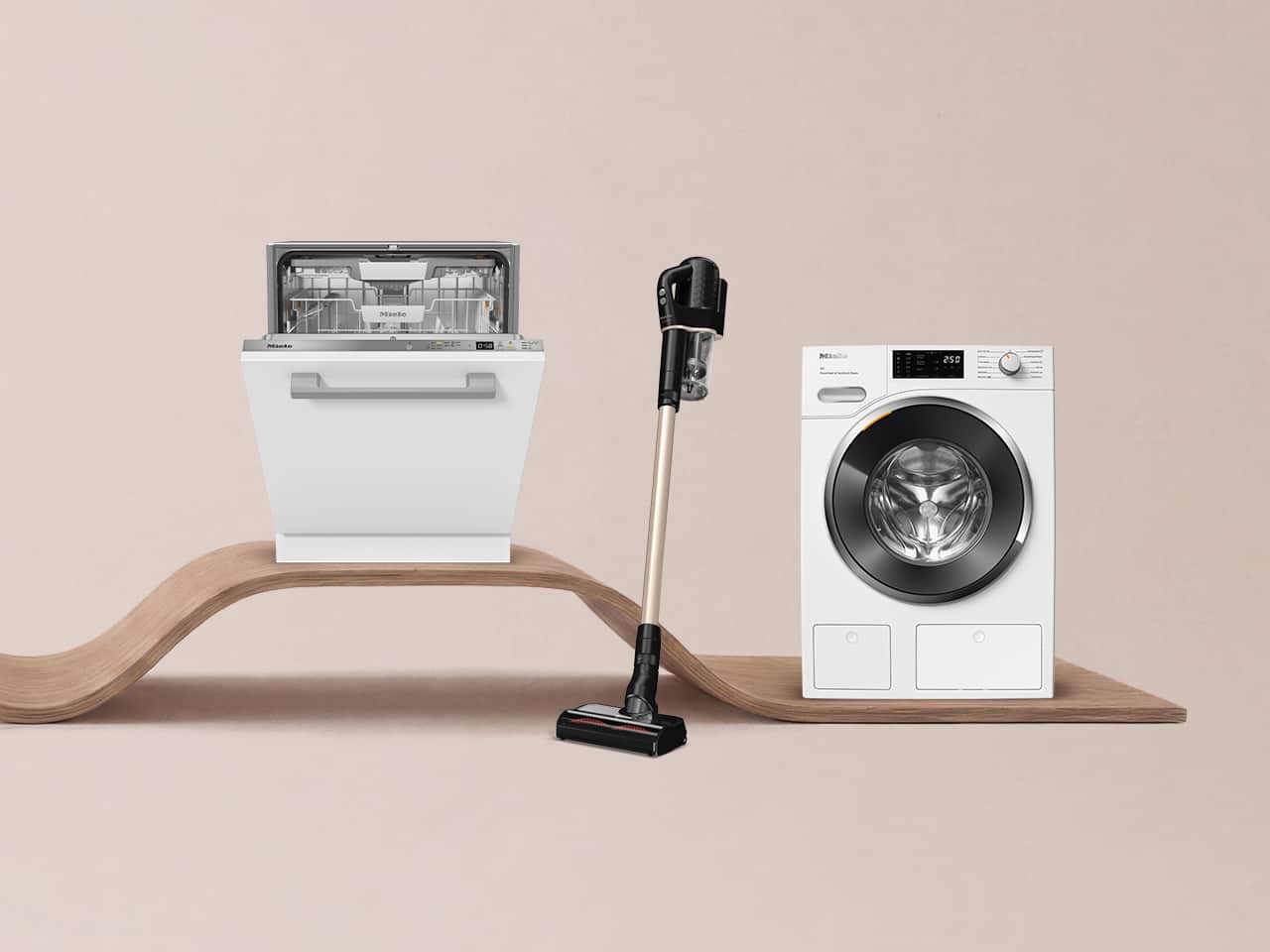 Dishwasher, cordless stick vacuum, and front-load washer displayed on a curved wooden shelf against a neutral backdrop.