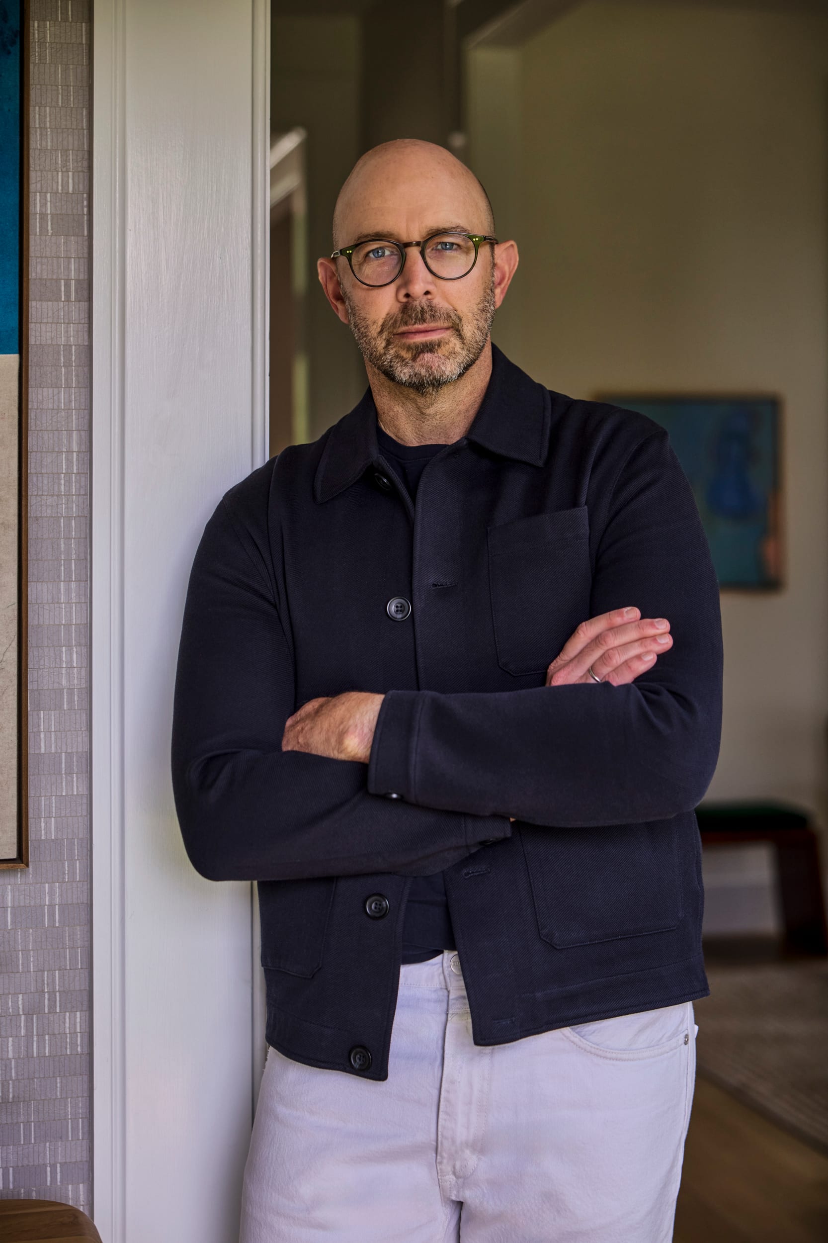 Portrait of a bald man with glasses, arms crossed in a dark jacket, standing indoors by a doorway; professional headshot.