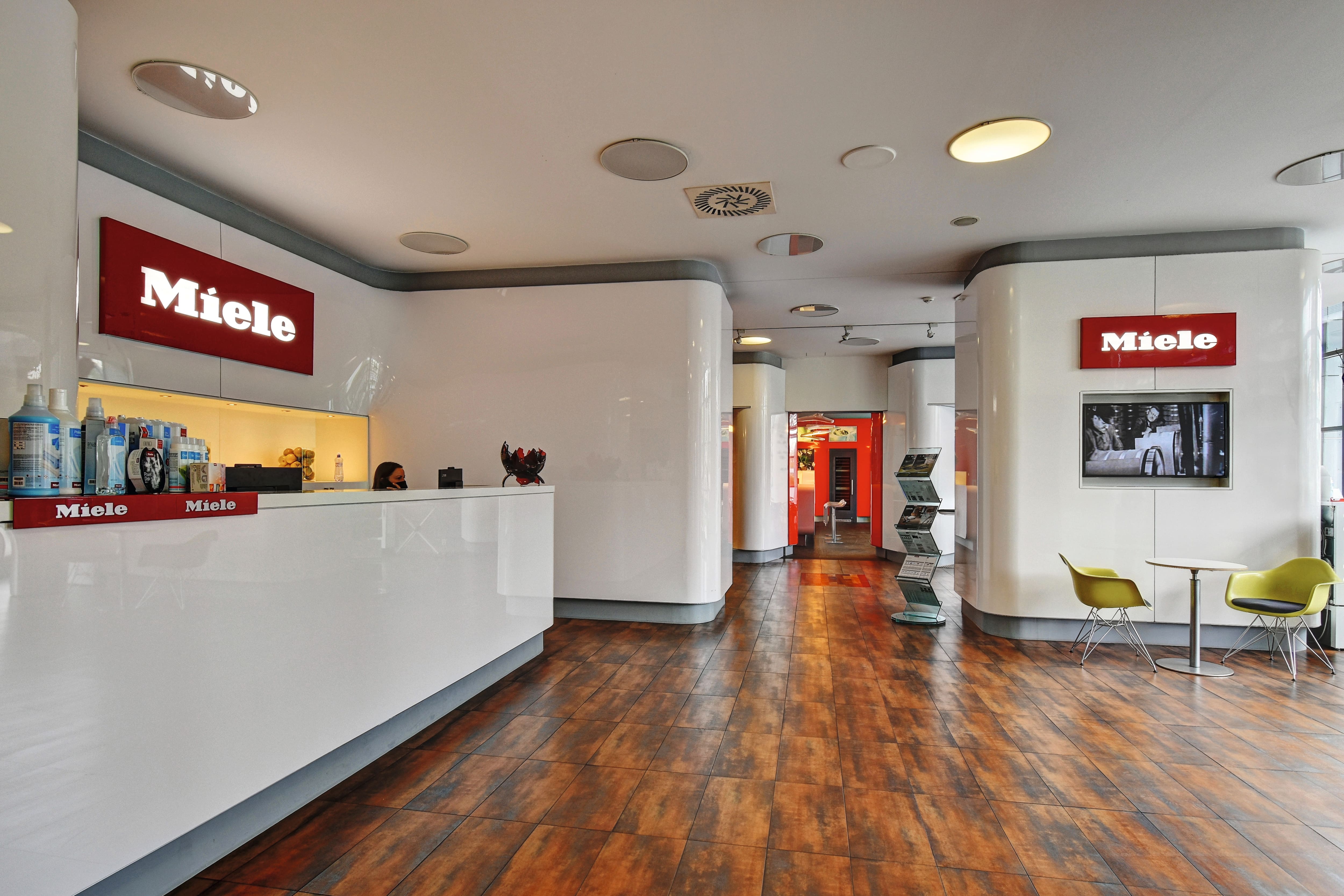 Moderní interiér showroomu s lesklými bílými stěnami a pulty, podlahou s dřevěným vzhledem, červenými nápisy Miele, recepčním pultem vlevo, dvěma zelenými židlemi a malým stolkem vpravo a chodbou vedoucí dále do obchodu. Moderní interiér showroomu s lesklými bílými stěnami a pulty, podlahou s dřevěným vzhledem, červenými nápisy Miele, recepčním pultem vlevo, dvěma zelenými židlemi a malým stolkem vpravo a chodbou vedoucí dále do obchodu.