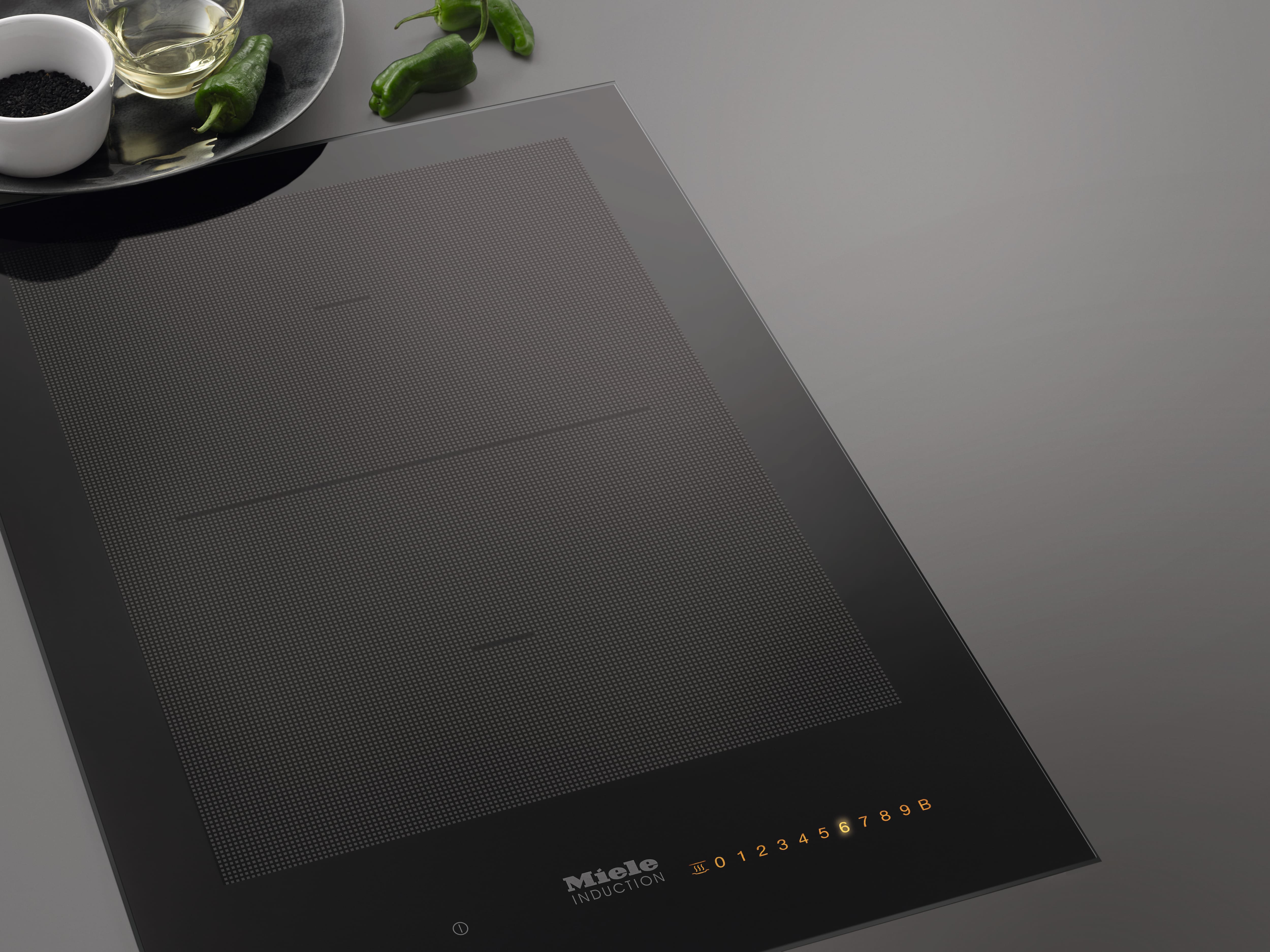 Close-up of a sleek black induction cooktop with glowing touch controls, beside peppers and oil on a plate. Close-up of a sleek black induction cooktop with glowing touch controls, beside peppers and oil on a plate.