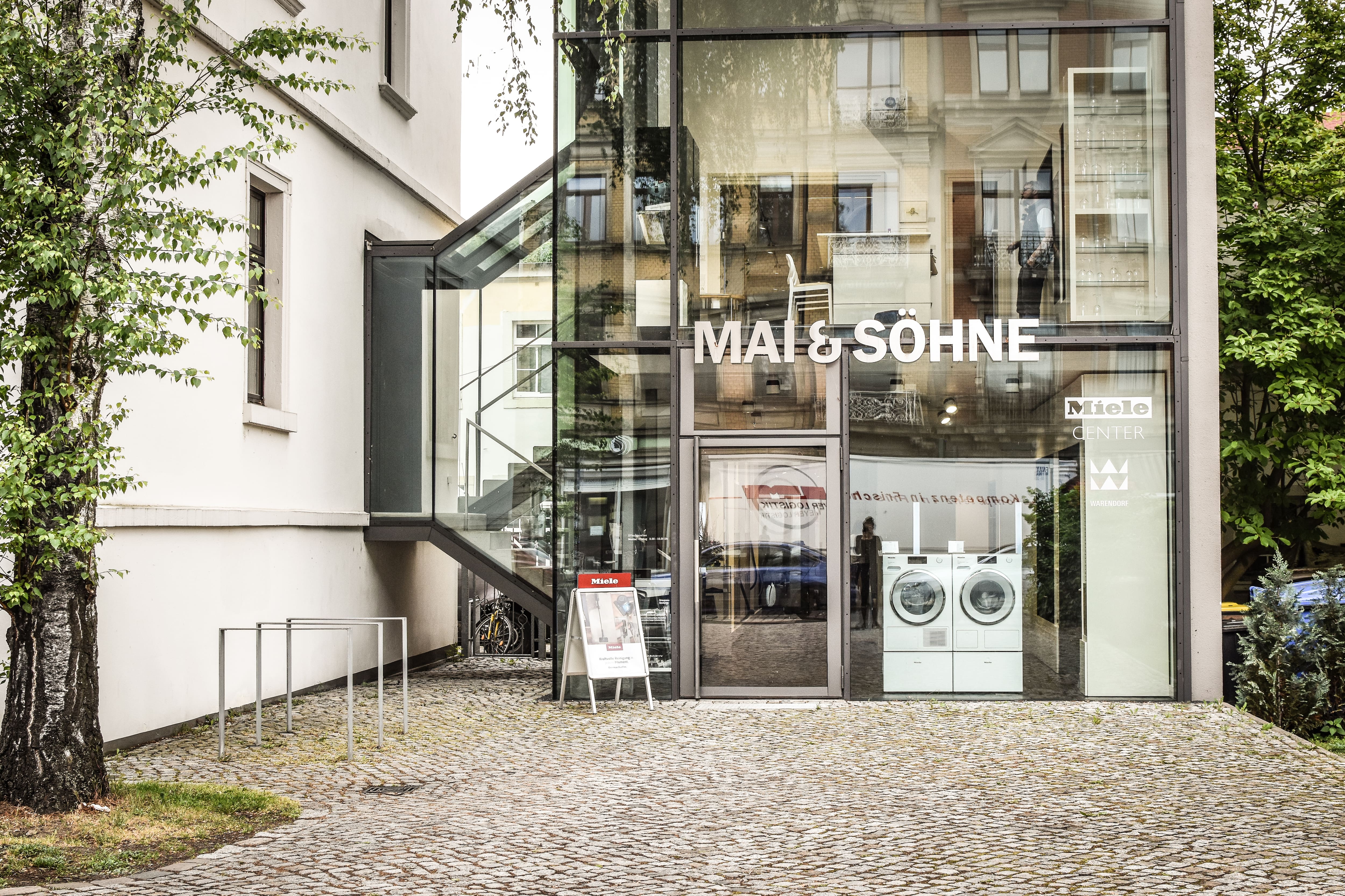 Glasfrontiges MAI & SÖHNE Geräte-Showroom mit Waschmaschinen im Schaufenster; Kopfsteinpflaster-Eingang und nahegelegene Bäume.