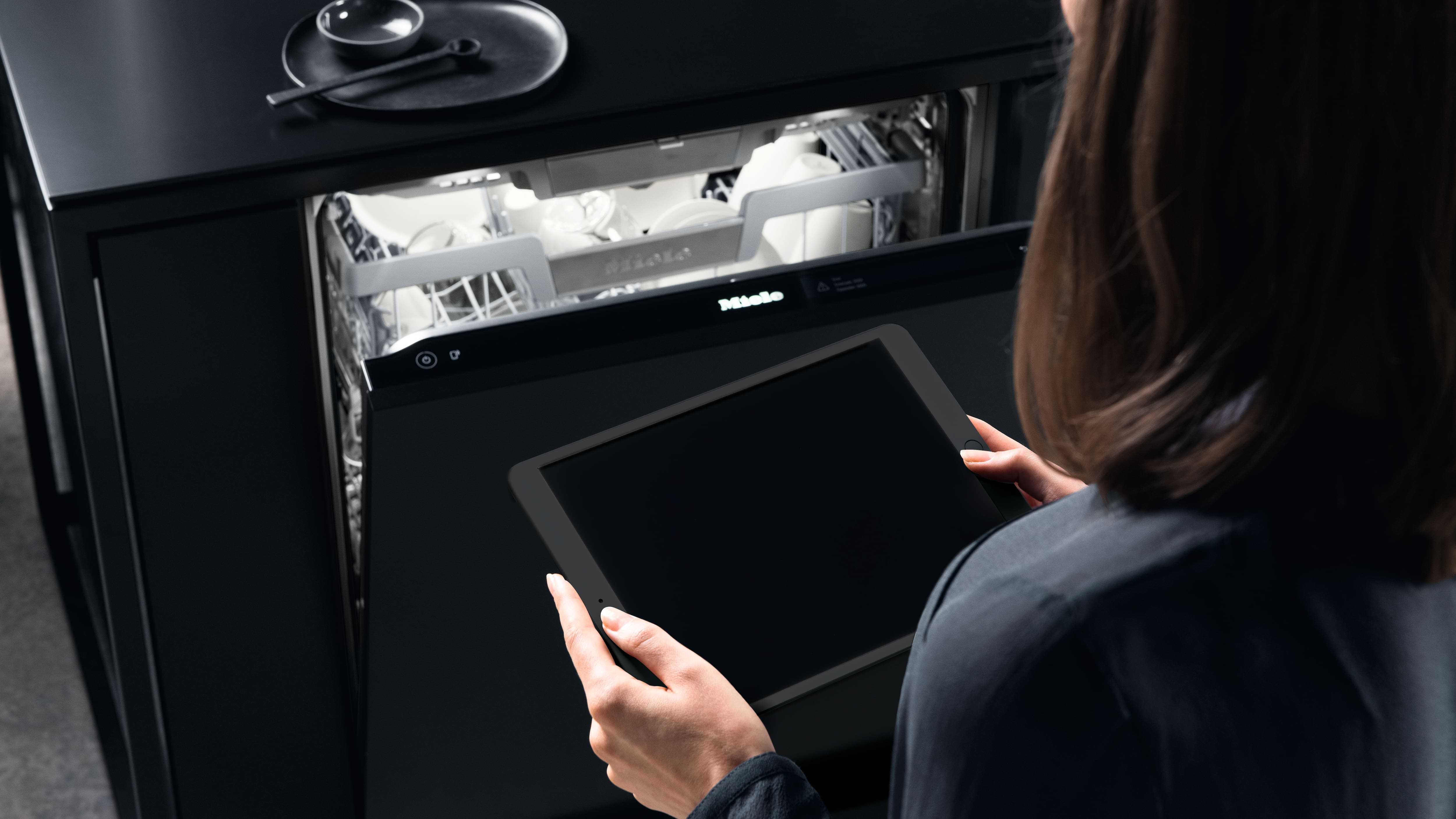 Person holding a tablet in a modern kitchen, standing by an open built-in dishwasher loaded with white dishes. Person holding a tablet in a modern kitchen, standing by an open built-in dishwasher loaded with white dishes.