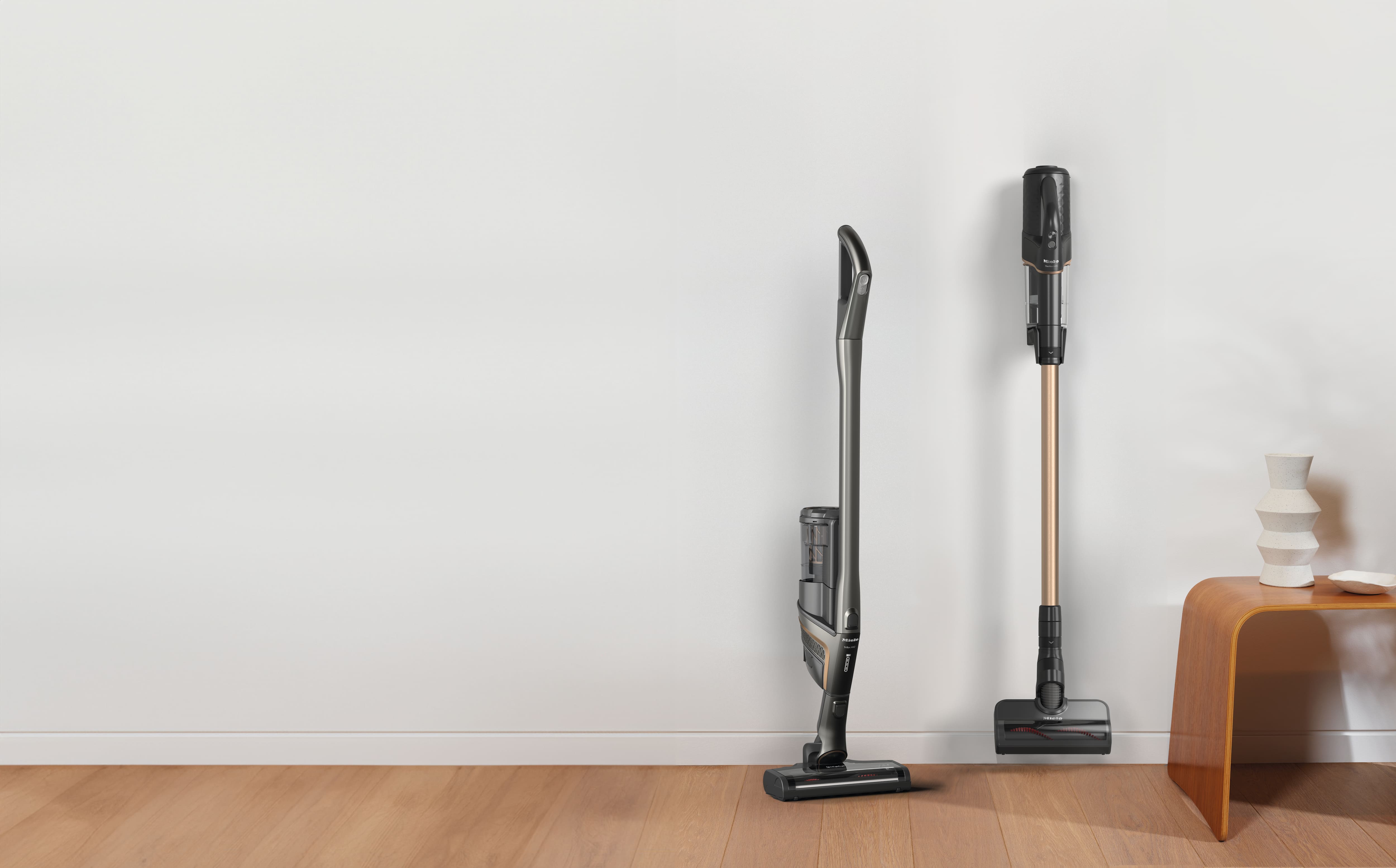 Zwei kabellose Stielstaubsauger stehen an einer weißen Wand auf einem Holzboden neben einer Holzbank mit Vasen. Zwei kabellose Stielstaubsauger stehen an einer weißen Wand auf einem Holzboden neben einer Holzbank mit Vasen.