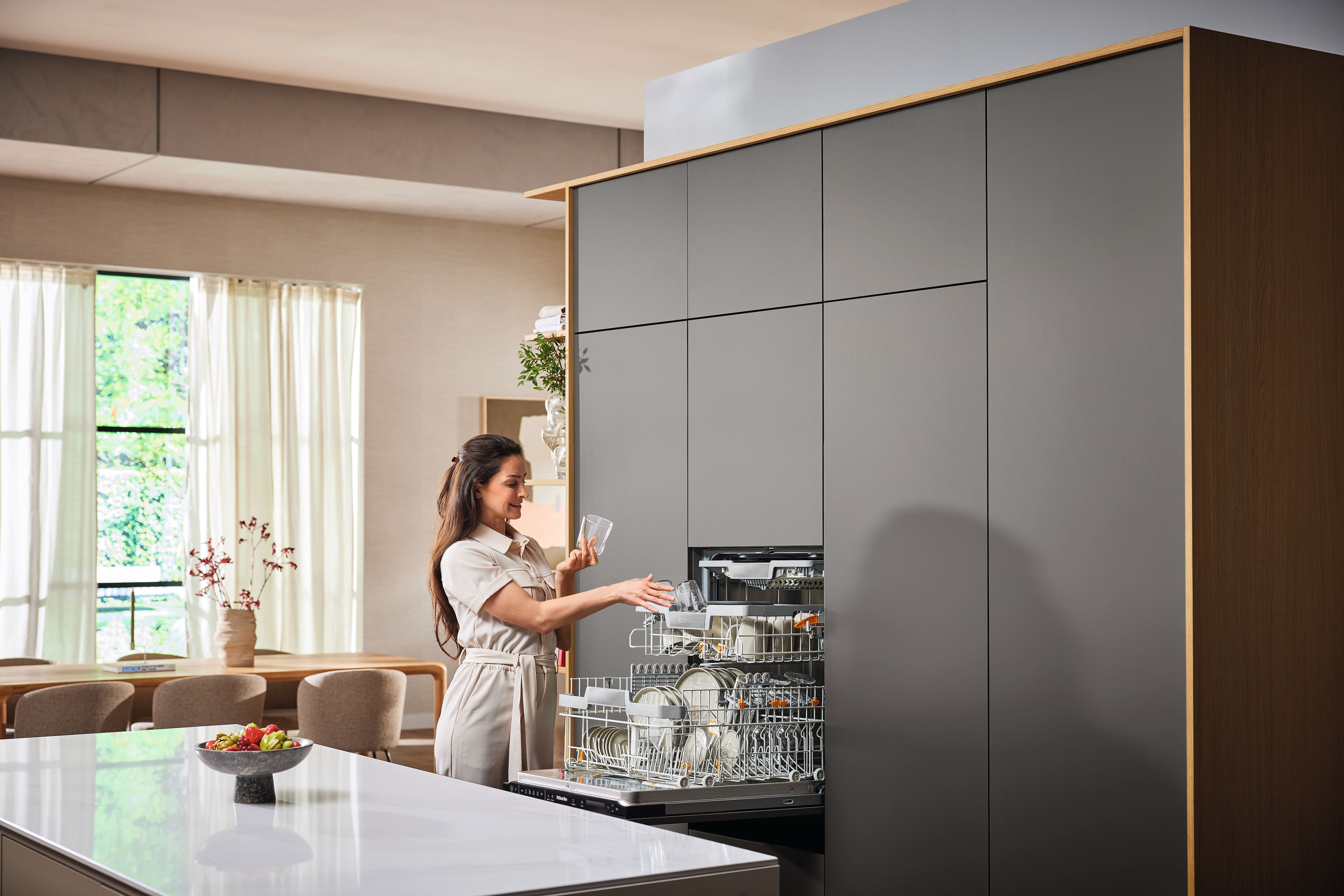 Person loading glasses into a built-in dishwasher in a modern, minimalist kitchen with gray cabinetry. Person loading glasses into a built-in dishwasher in a modern, minimalist kitchen with gray cabinetry.