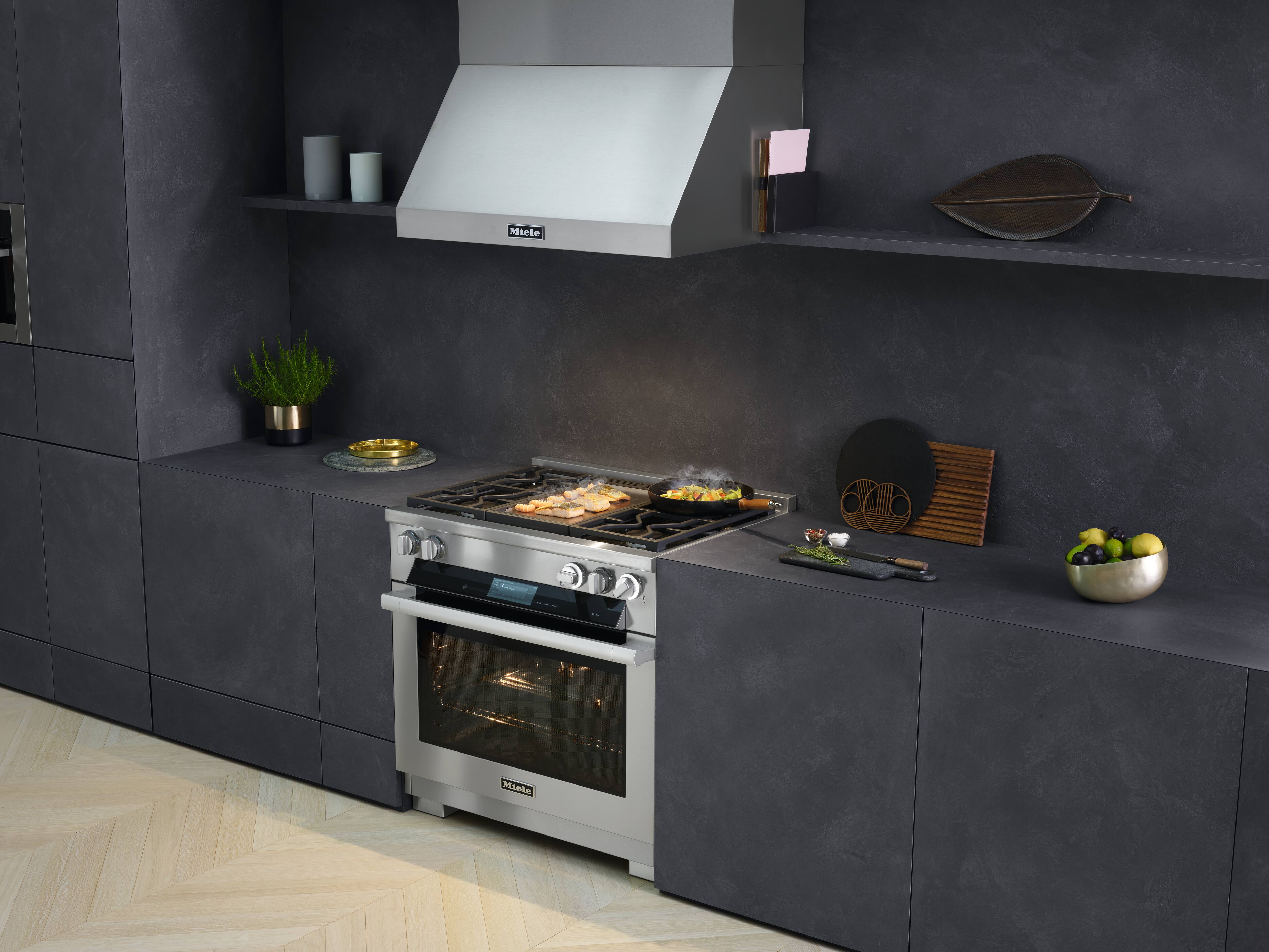 Cozinha moderna em tons escuros, com fogão e exaustor de aço inoxidável; alimentos chiando no fogão a gás; ervas e utensílios por perto. Cozinha moderna em tons escuros, com fogão e exaustor de aço inoxidável; alimentos chiando no fogão a gás; ervas e utensílios por perto.