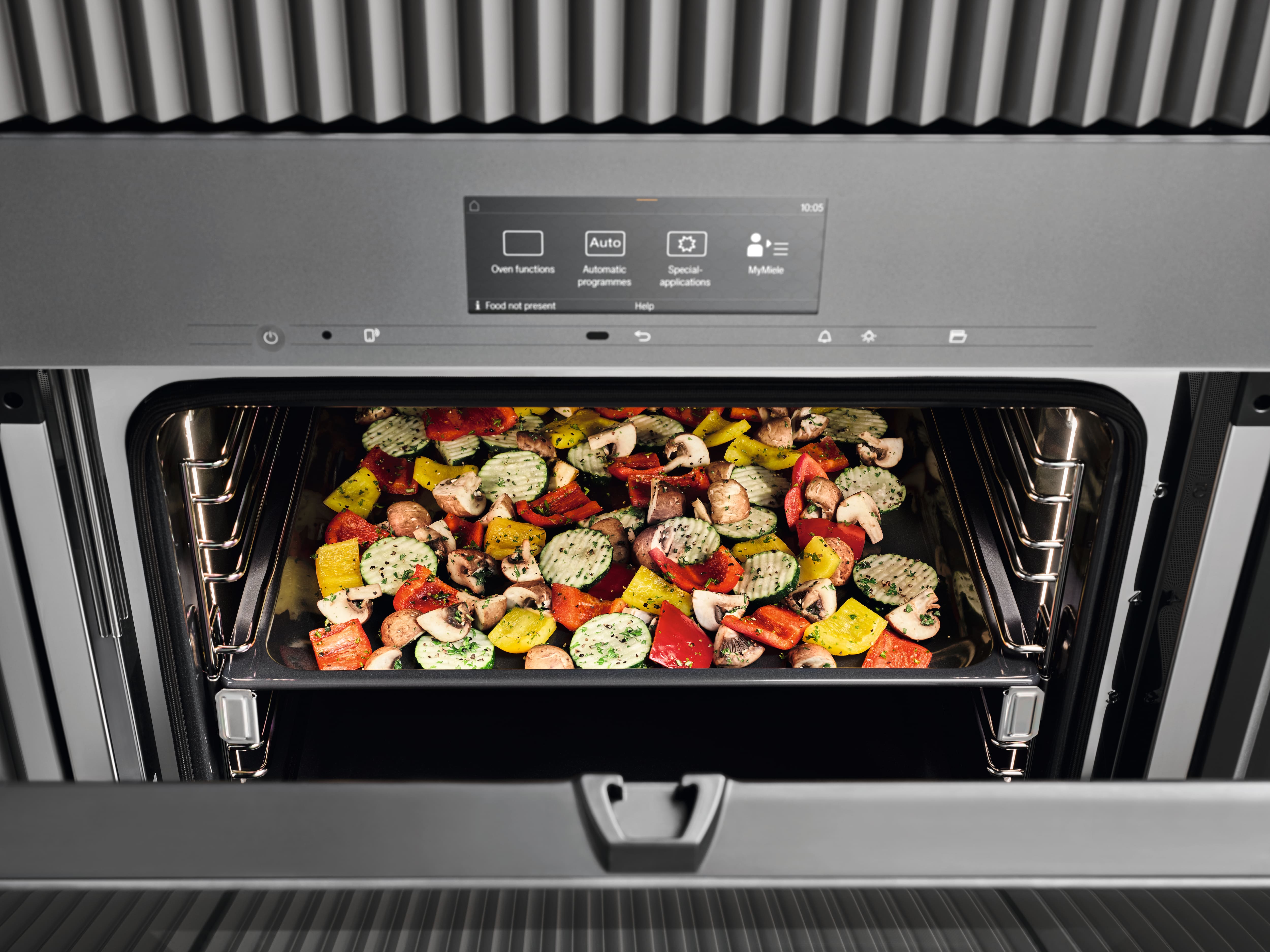 Open modern oven with a touchscreen control panel; a baking tray of colorful chopped vegetables—peppers, mushrooms, and zucchini—is roasting inside.