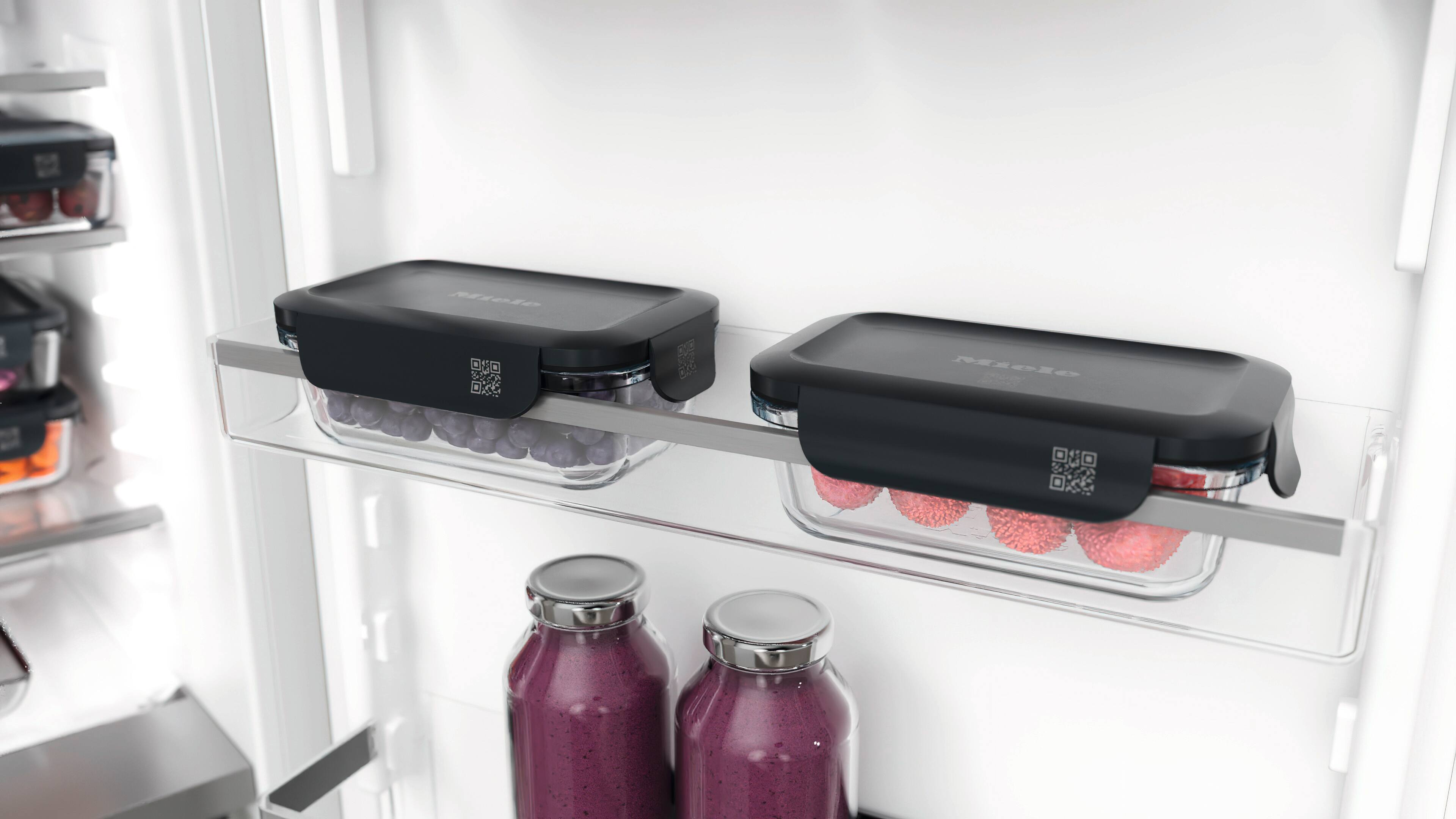 Inside a refrigerator, two rectangular glass food containers with black lids and QR code labels sit on a shelf—one filled with purple grapes and the other with pink berries—above two bottles of purple smoothie. Inside a refrigerator, two rectangular glass food containers with black lids and QR code labels sit on a shelf—one filled with purple grapes and the other with pink berries—above two bottles of purple smoothie.