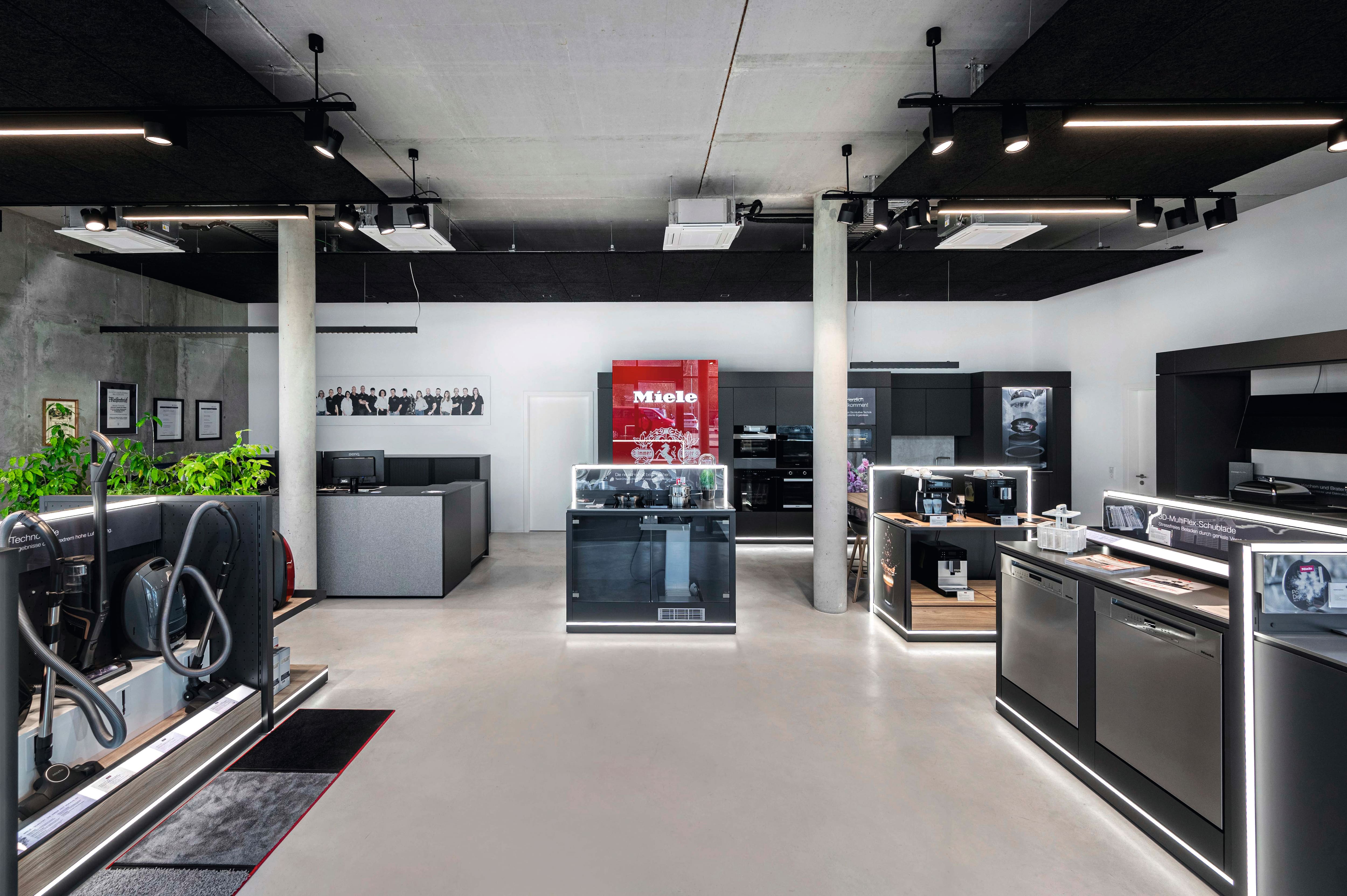 Moderner Geräteschauraum mit Miele-Branding, mit eleganten schwarzen Displays von Geschirrspülern, Kaffeemaschinen und Staubsaugern unter Schienenbeleuchtung und Betonpfeilern.