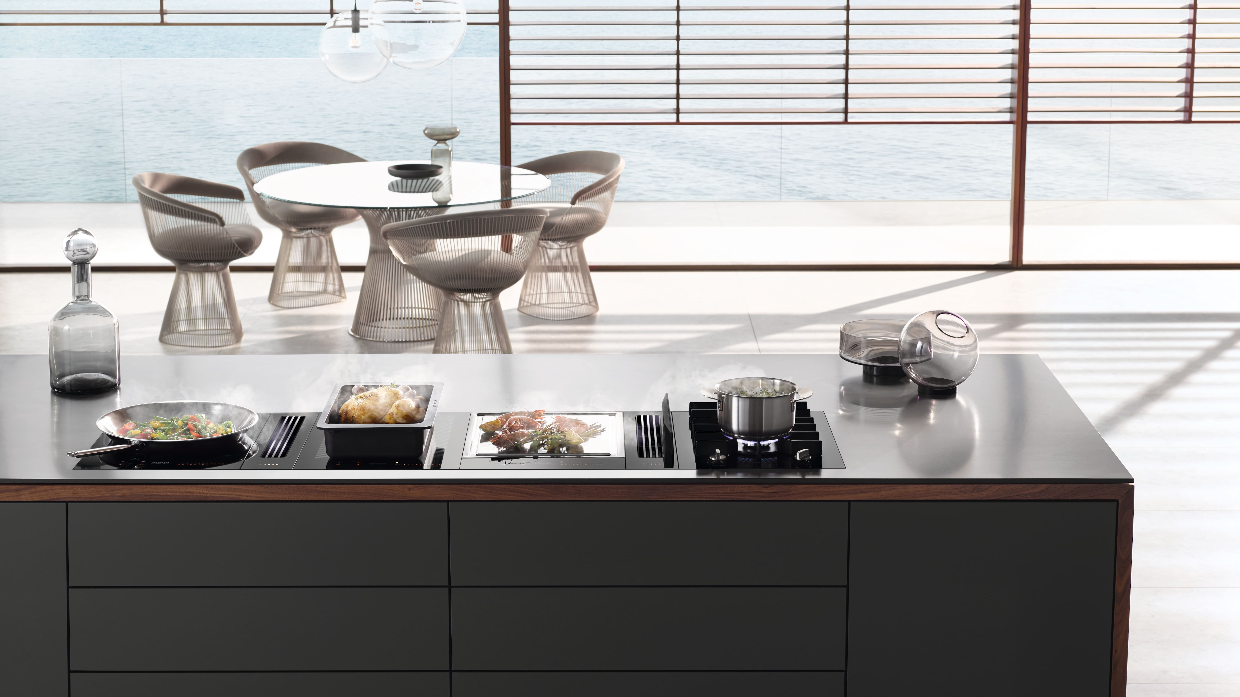 Modern kitchen island with modular cooktops cooking meals, overlooking a glass-walled dining area by the sea. Modern kitchen island with modular cooktops cooking meals, overlooking a glass-walled dining area by the sea.