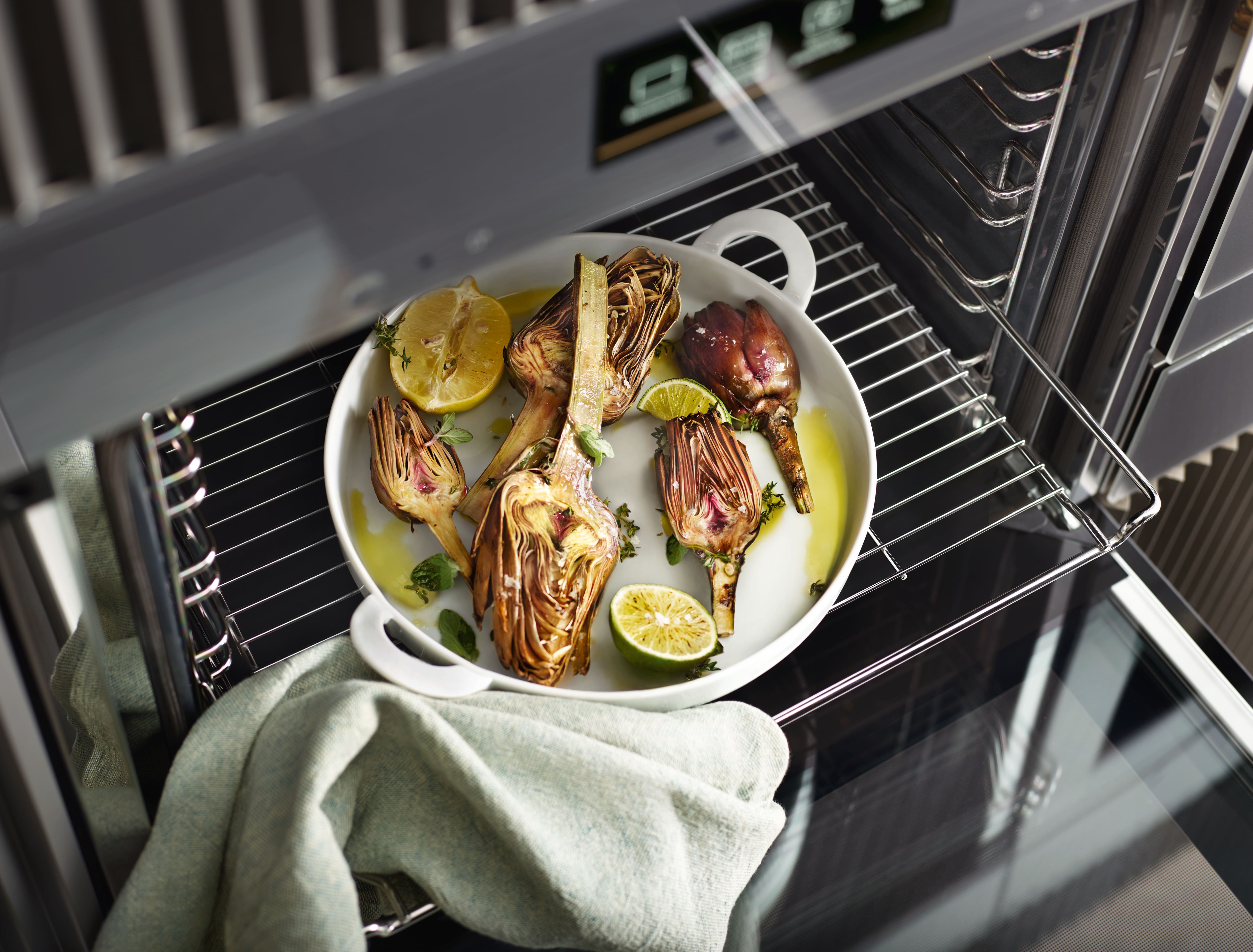 White baking dish of roasted halved artichokes with lemon and lime slices and herbs on an oven rack, with a green kitchen towel draped nearby.