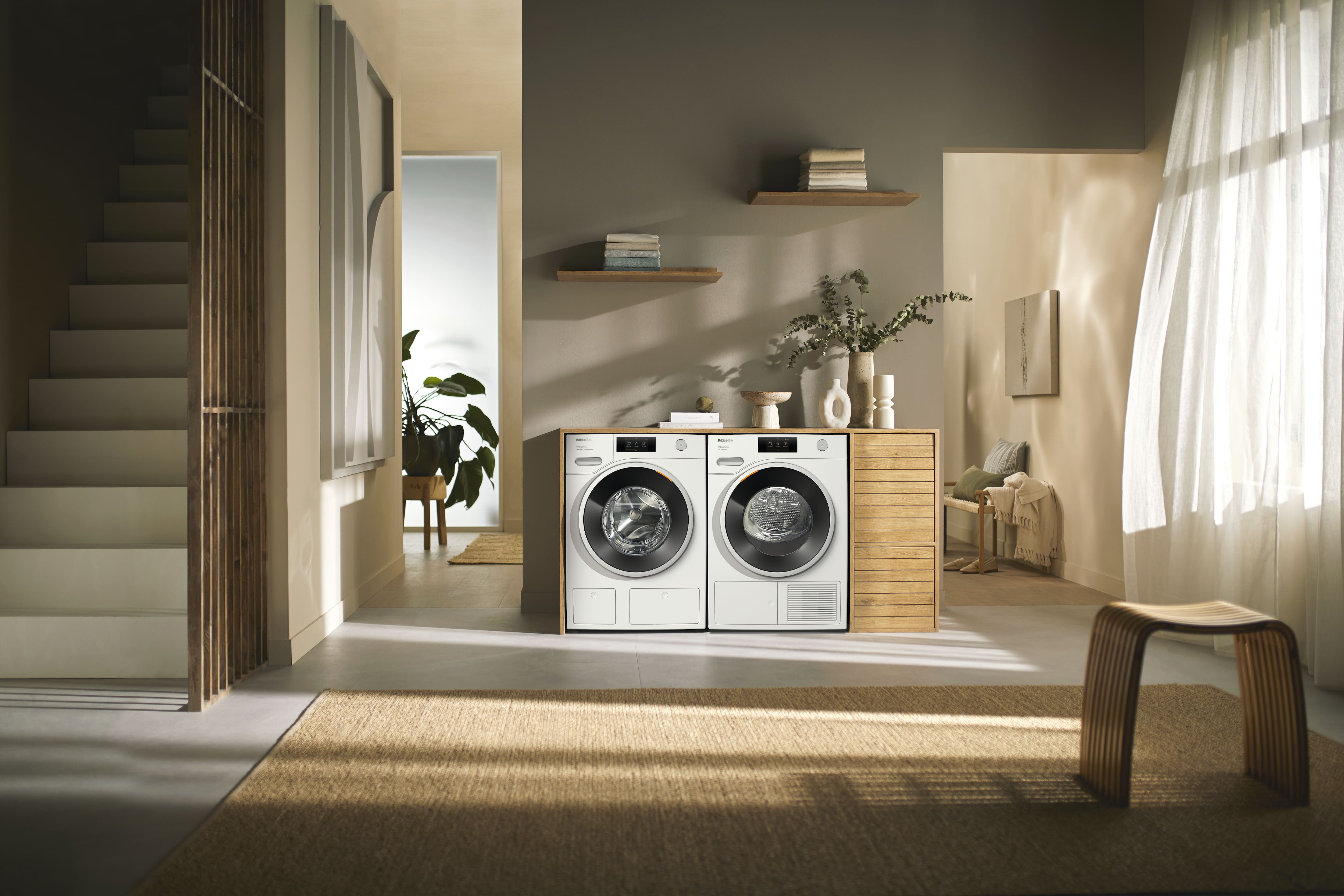 Modern laundry nook with side-by-side front-load washer and dryer in wood cabinet, sunlit room with shelf, plant and stool. Modern laundry nook with side-by-side front-load washer and dryer in wood cabinet, sunlit room with shelf, plant and stool.