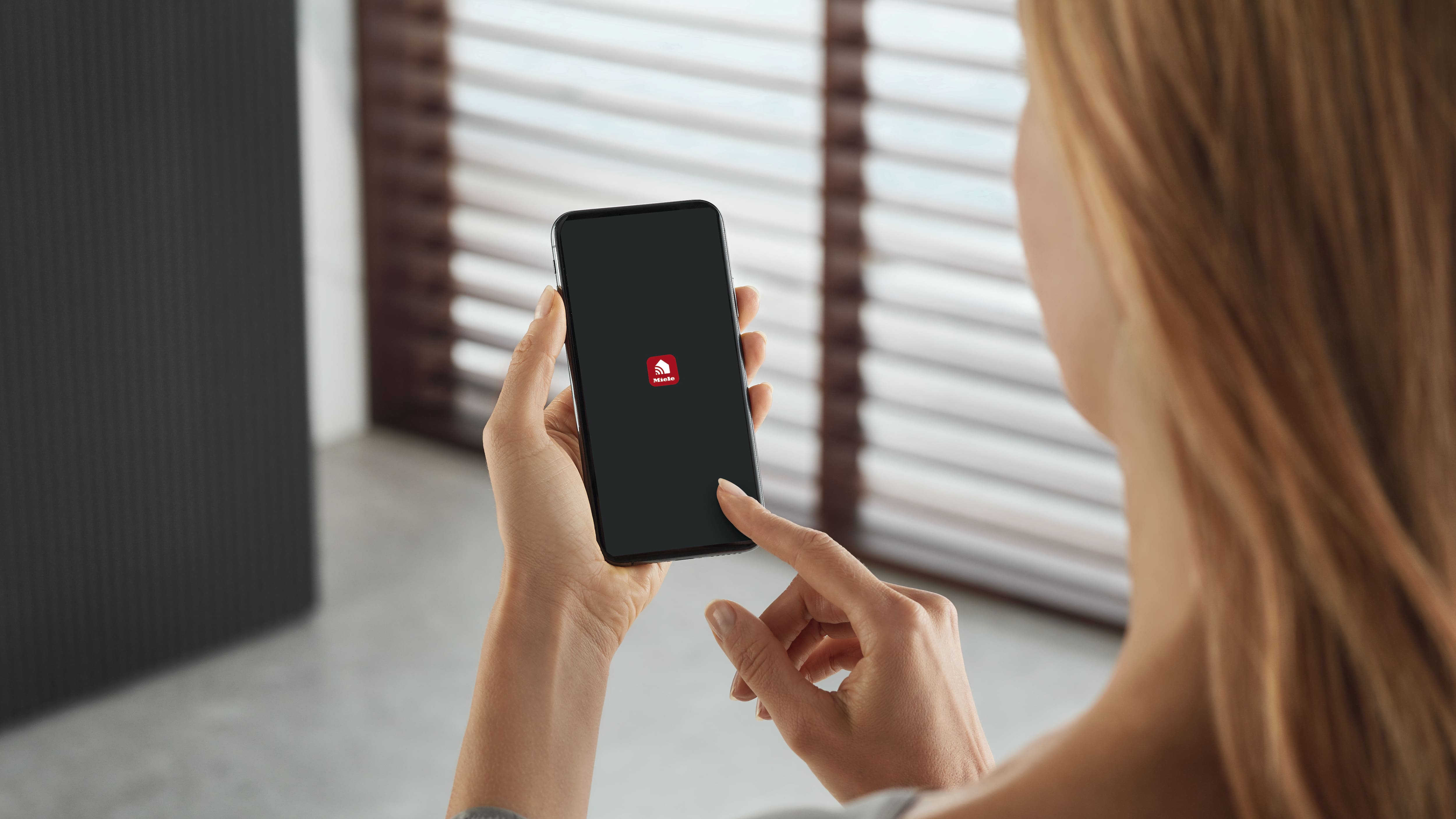 Pessoa segurando um smartphone e tocando para abrir a app Miele, sugerindo controlo de eletrodomésticos inteligentes. Pessoa segurando um smartphone e tocando para abrir a app Miele, sugerindo controlo de eletrodomésticos inteligentes.