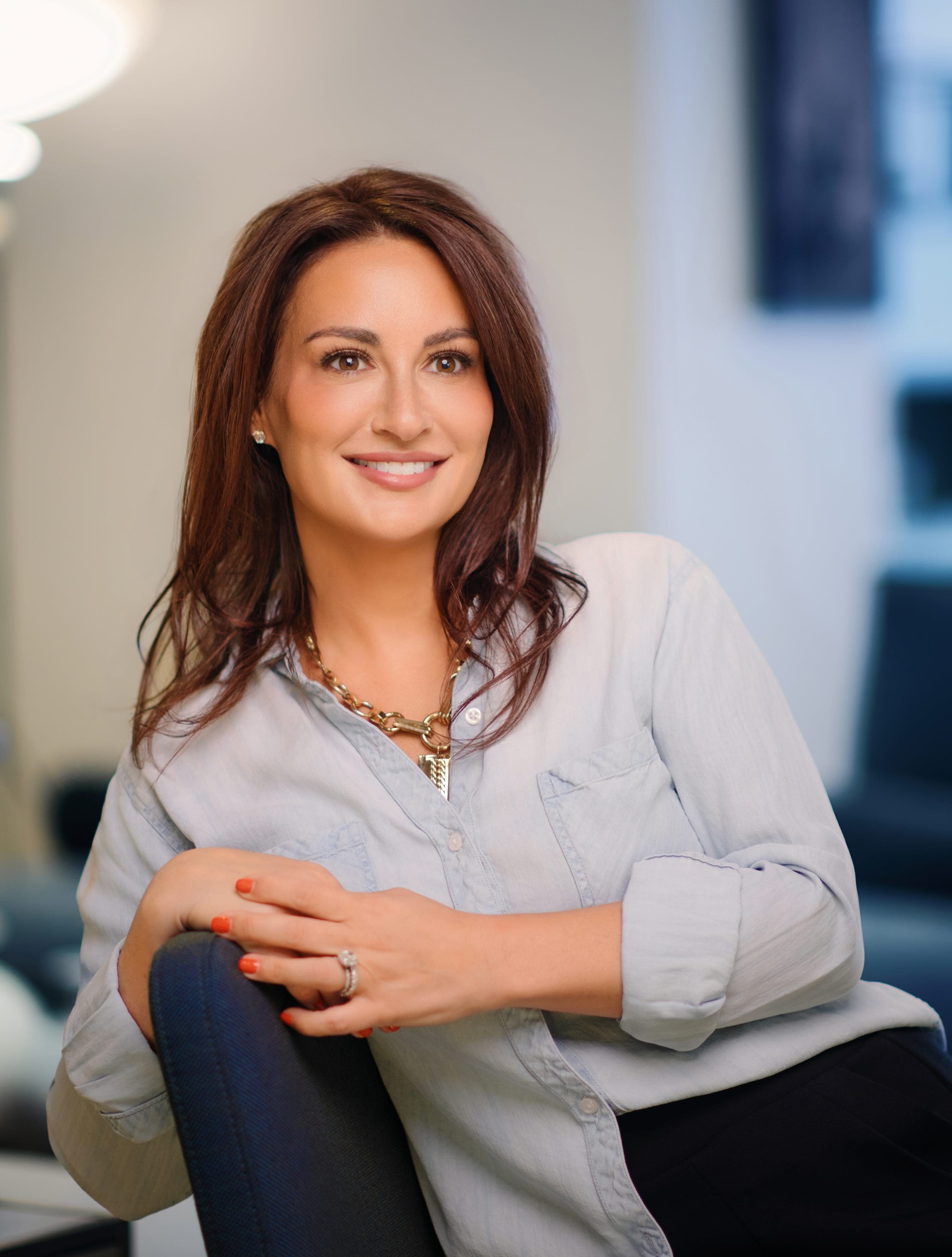 Professional headshot of a smiling woman in a light button-up blouse, leaning on a chair in a modern office.