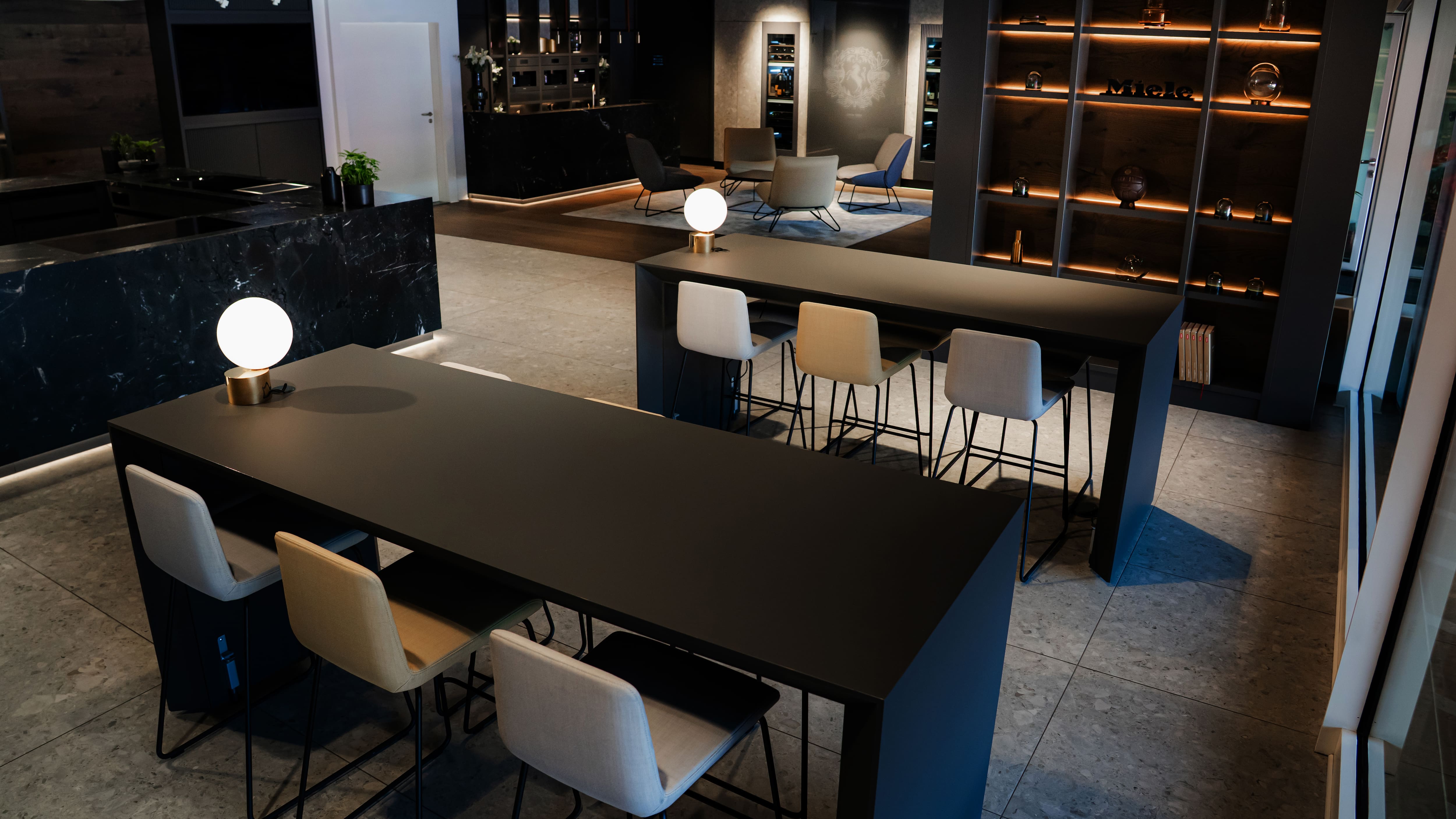 Moderne Lounge mit eleganten schwarzen Tischen, Barhockern, kugelförmigen Pendelleuchten und indirekt beleuchteten Regalen. Das stilvolle Showroom-Ambiente vermittelt ein hochwertiges und zeitgemäßes Design. Moderne Lounge mit eleganten schwarzen Tischen, Barhockern, kugelförmigen Pendelleuchten und indirekt beleuchteten Regalen. Das stilvolle Showroom-Ambiente vermittelt ein hochwertiges und zeitgemäßes Design.