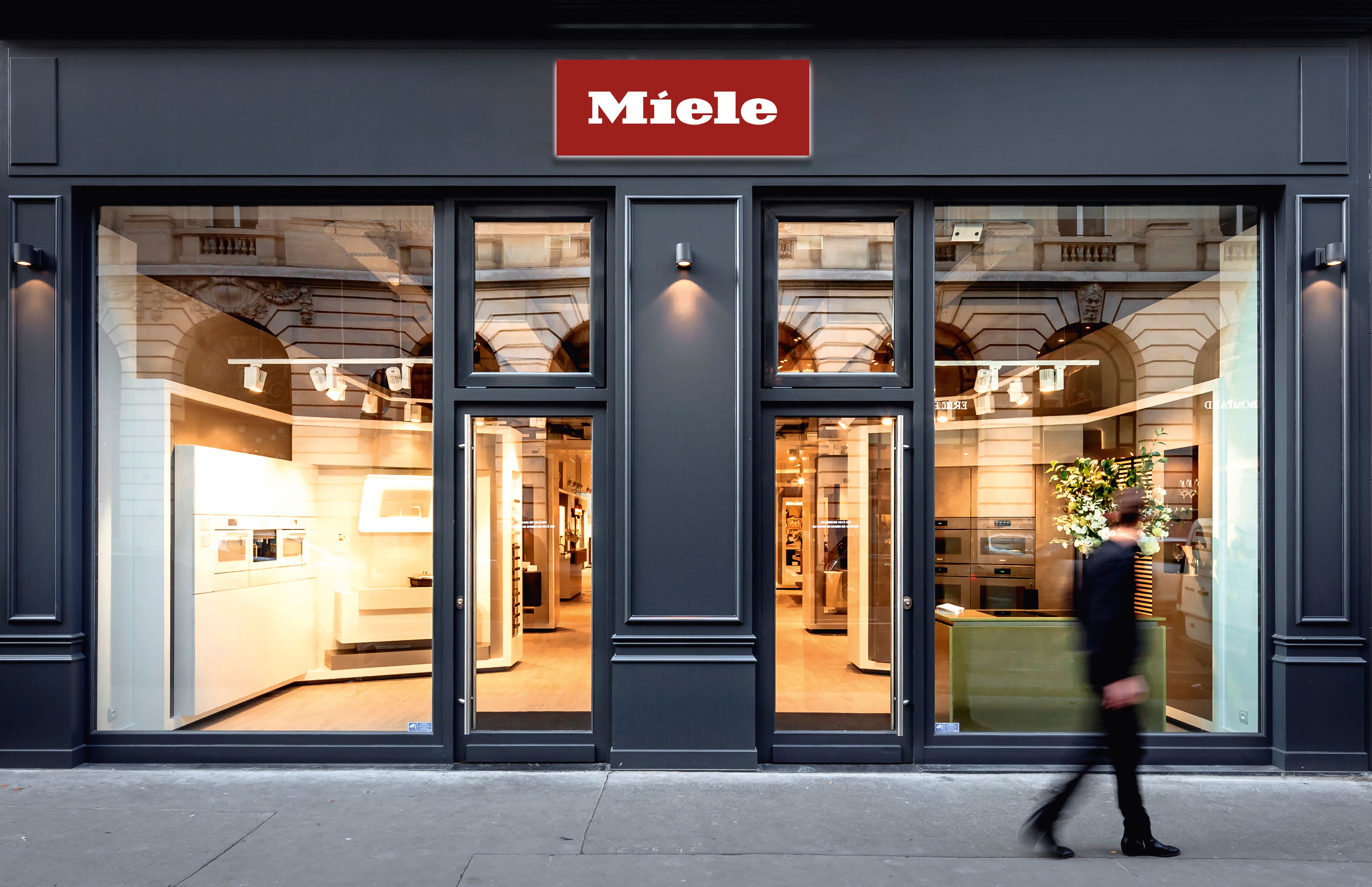 Vue extérieure d'un magasin Miele moderne, avec de grandes baies vitrées, un éclairage intérieur chaleureux et un passant. Vue extérieure d'un magasin Miele moderne, avec de grandes baies vitrées, un éclairage intérieur chaleureux et un passant.