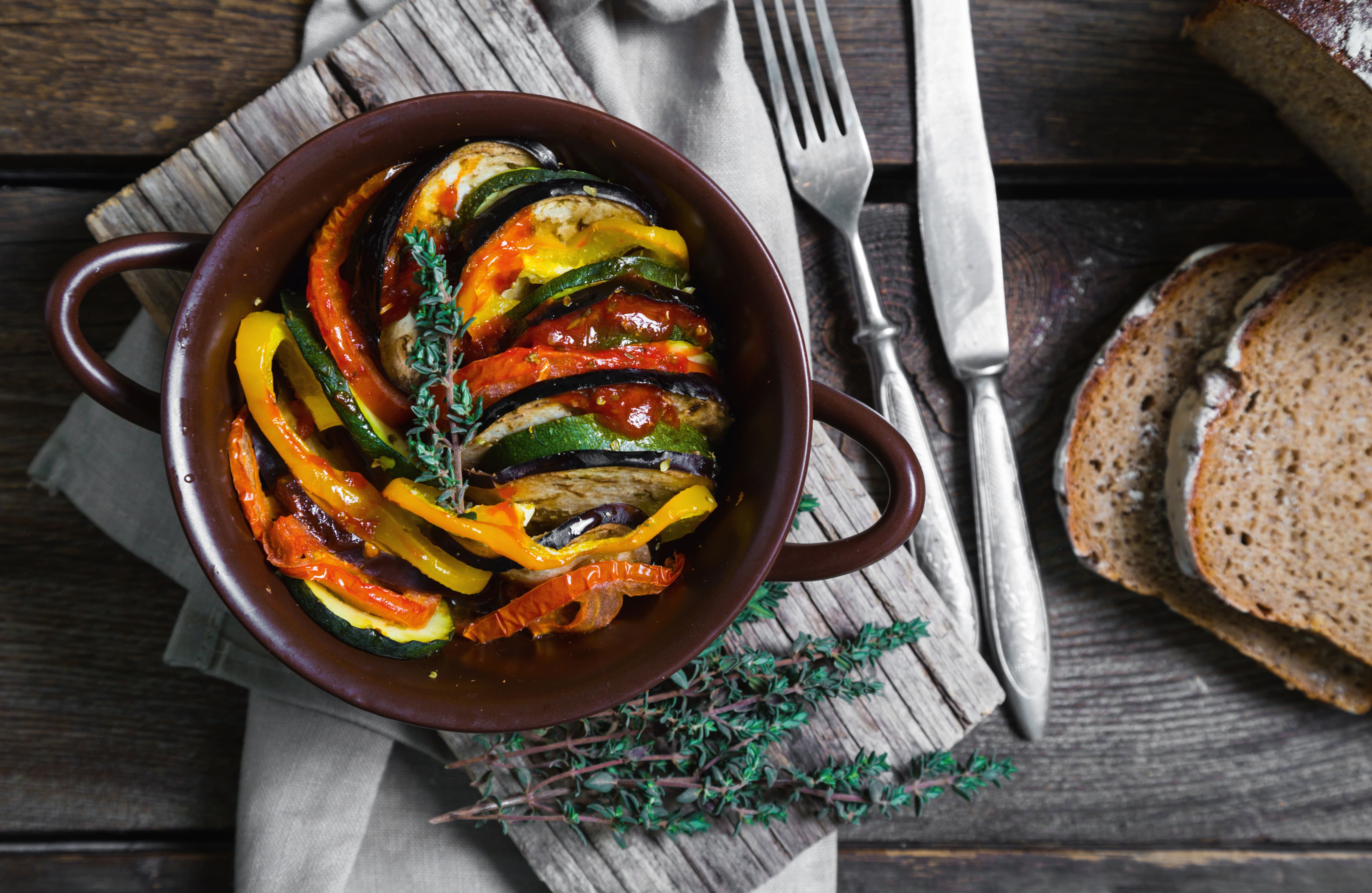 Top-down view of ratatouille in a brown casserole, layered eggplant, zucchini, peppers, with thyme; bread and cutlery. Top-down view of ratatouille in a brown casserole, layered eggplant, zucchini, peppers, with thyme; bread and cutlery.