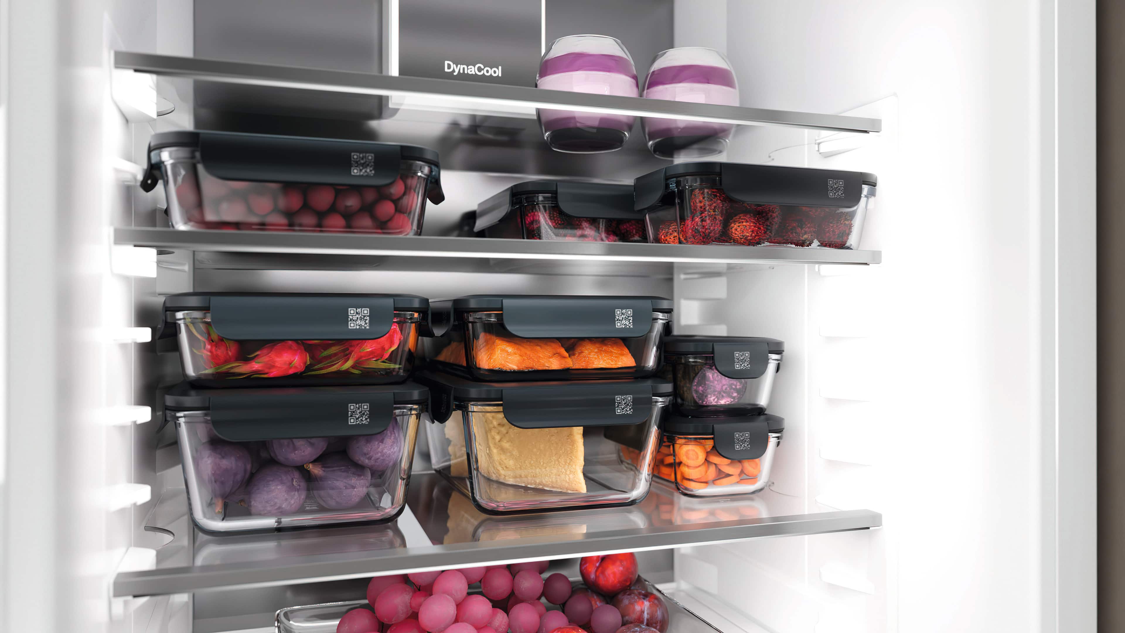 Open refrigerator interior with glass shelves holding multiple clear containers with dark lids, filled with assorted foods such as cherries or grapes, lychees, dragon fruit, purple onions, sliced carrots, salmon fillets, and a block of cheese, plus two jars of layered yogurt on the top shelf; “DynaCool” text visible on the back panel. Open refrigerator interior with glass shelves holding multiple clear containers with dark lids, filled with assorted foods such as cherries or grapes, lychees, dragon fruit, purple onions, sliced carrots, salmon fillets, and a block of cheese, plus two jars of layered yogurt on the top shelf; “DynaCool” text visible on the back panel.