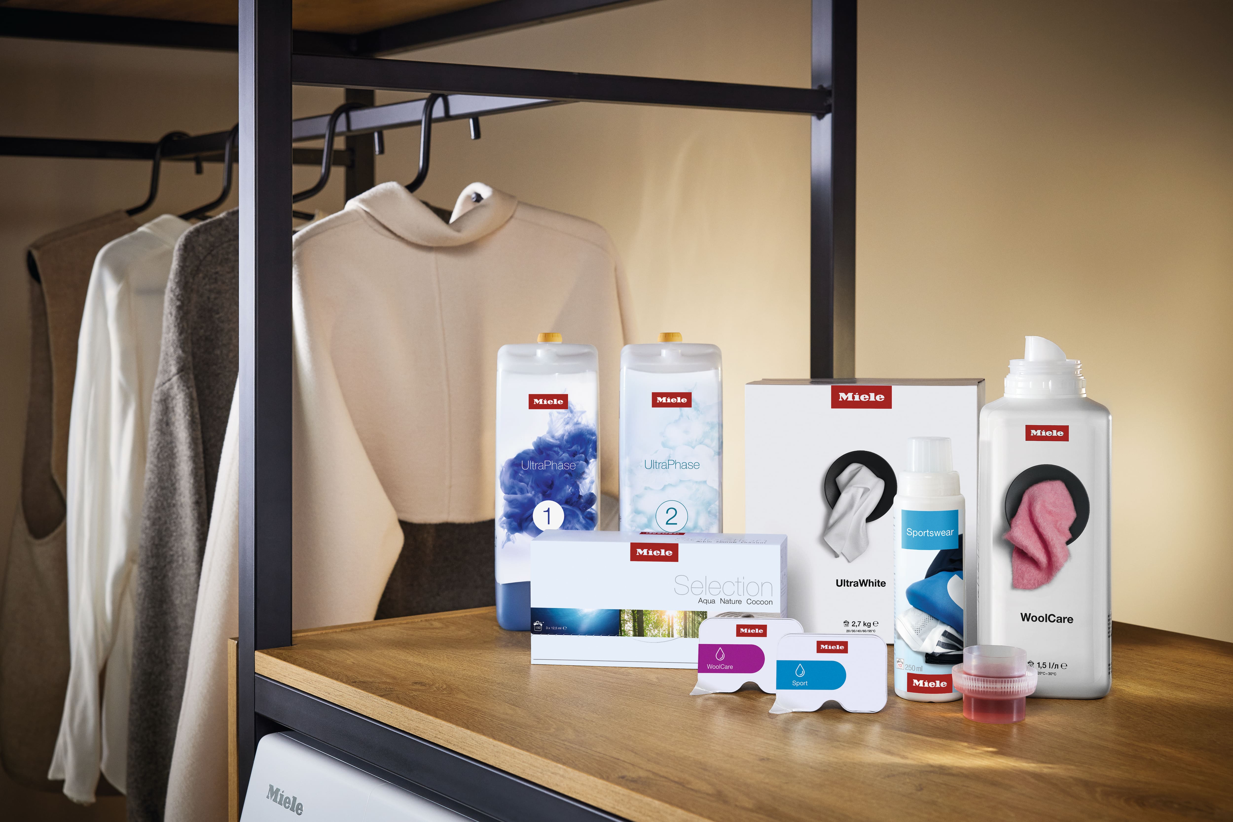 Selezione di prodotti Miele per il lavaggio e la cura dei tessuti – tra cui le cartucce UltraPhase 1 e 2, UltraWhite, WoolCare, Sportswear e Caps – su una mensola in legno accanto a capi appesi. Selezione di prodotti Miele per il lavaggio e la cura dei tessuti – tra cui le cartucce UltraPhase 1 e 2, UltraWhite, WoolCare, Sportswear e Caps – su una mensola in legno accanto a capi appesi.