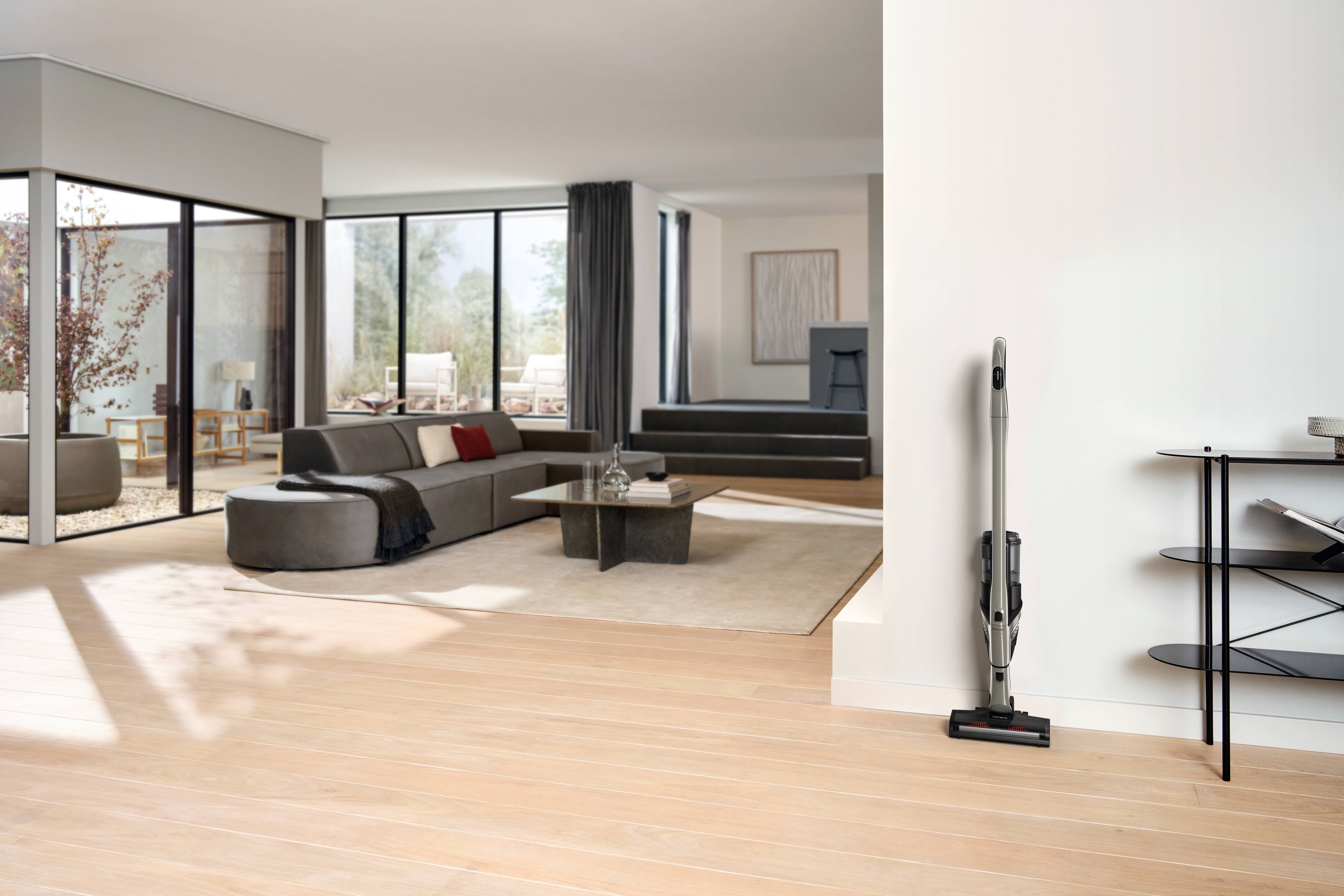 Moderne Wohnzimmer mit großen Fenstern, grauem Ecksofa und einem kabellosen Staubsauger, der an einer weißen Wand angedockt ist. Moderne Wohnzimmer mit großen Fenstern, grauem Ecksofa und einem kabellosen Staubsauger, der an einer weißen Wand angedockt ist.