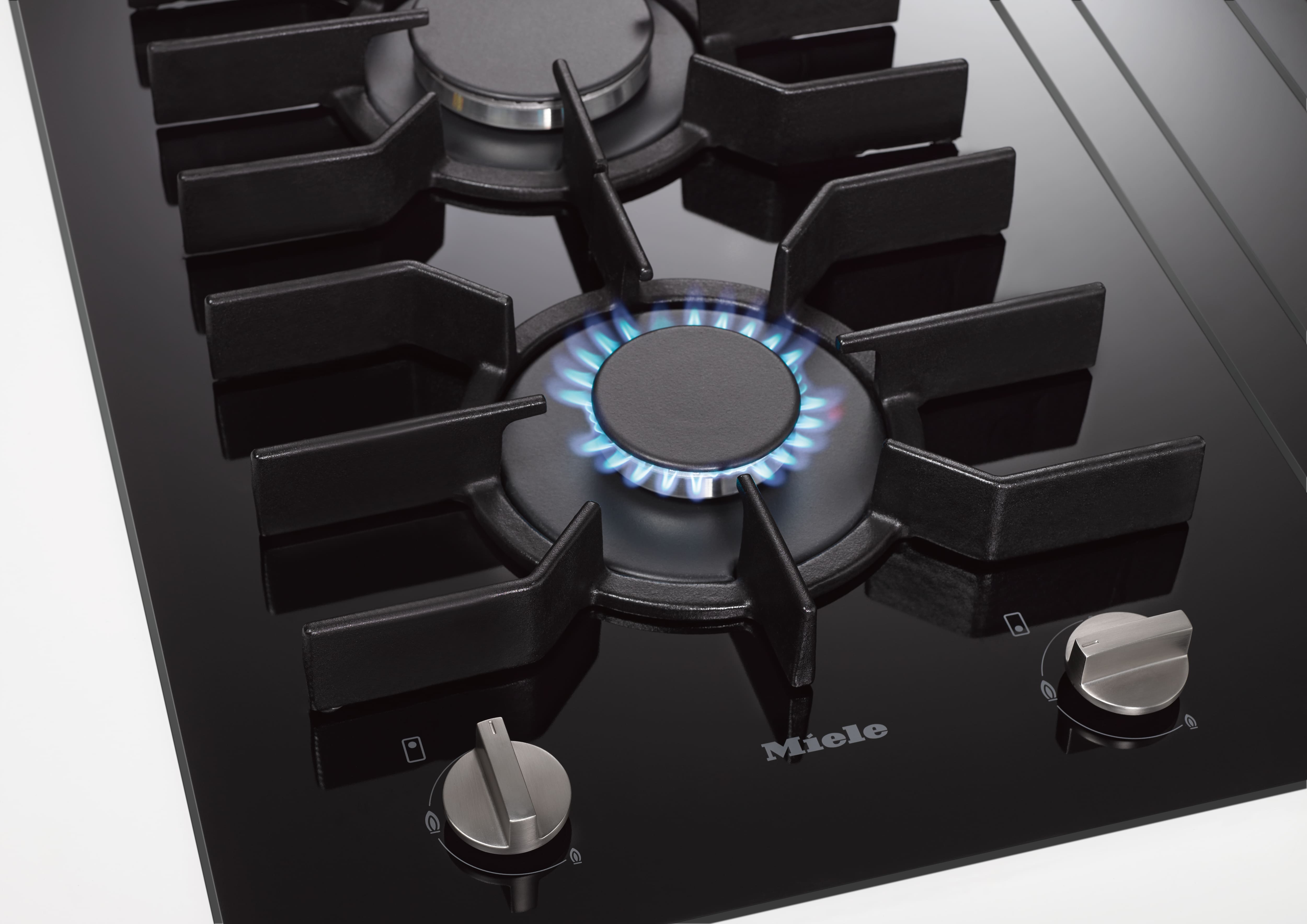 Close-up of a sleek black glass Miele gas cooktop with cast-iron grate, brass burner, and two control knobs; a small bowl of grains with a spoon sits on the counter beside it. Close-up of a sleek black glass Miele gas cooktop with cast-iron grate, brass burner, and two control knobs; a small bowl of grains with a spoon sits on the counter beside it.