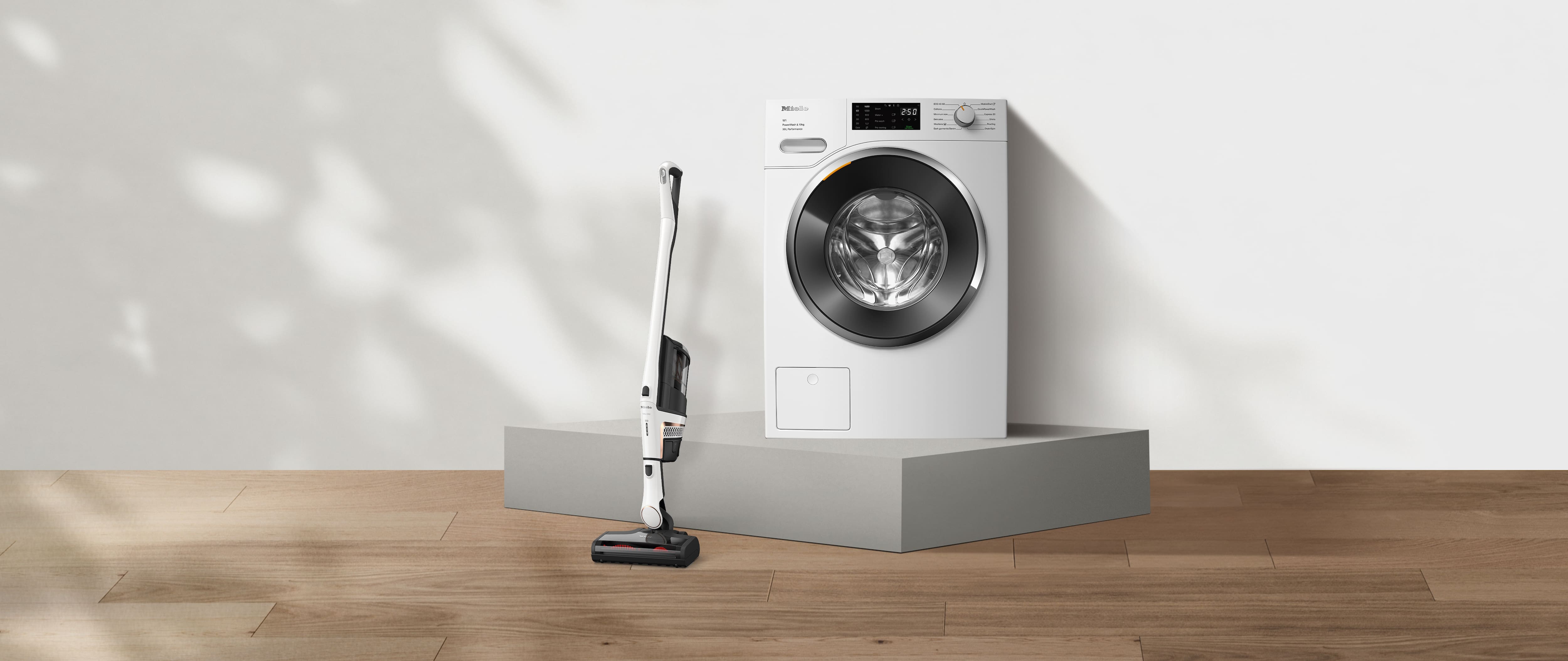 Minimalist scene with a white front-load washing machine on a pedestal and a cordless stick vacuum on wood flooring, soft natural light.
