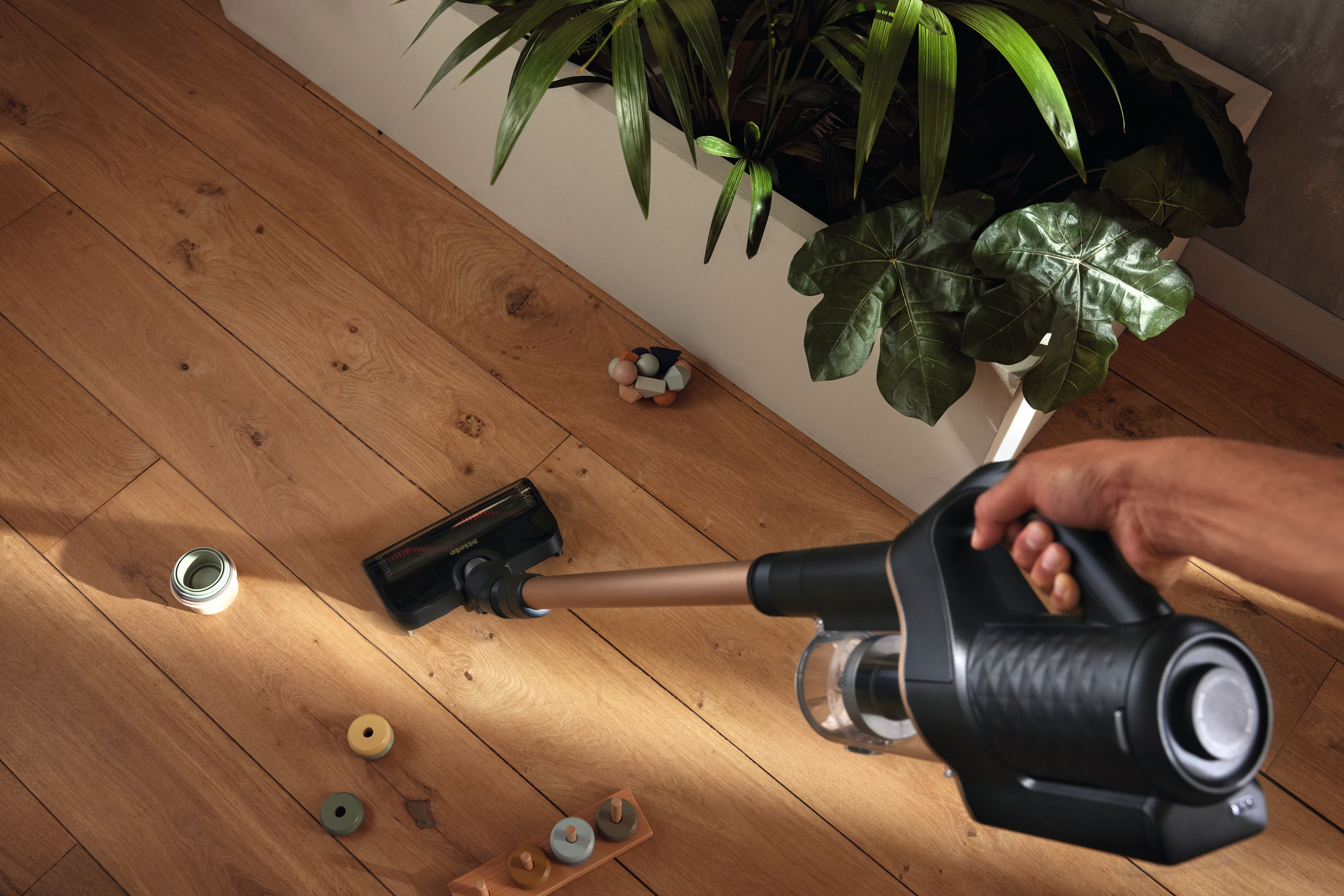 Hand using a cordless stick vacuum to clean a wooden floor near an indoor planter, with small toys scattered on the floor. Hand using a cordless stick vacuum to clean a wooden floor near an indoor planter, with small toys scattered on the floor.