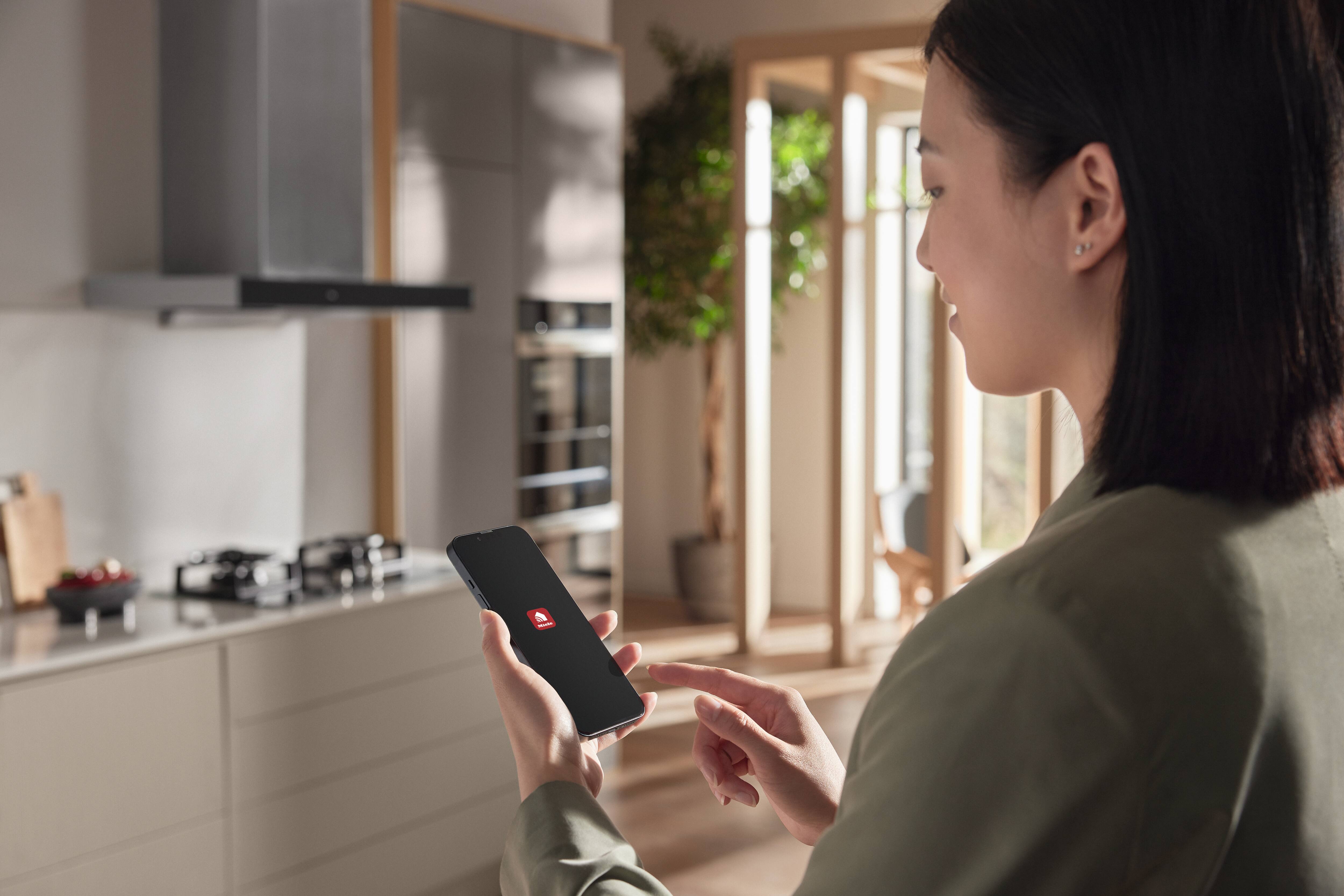 Uma pessoa em uma cozinha moderna usa um aplicativo de smartphone com um ícone de casa para controlar eletrodomésticos inteligentes e as configurações da casa. Uma pessoa em uma cozinha moderna usa um aplicativo de smartphone com um ícone de casa para controlar eletrodomésticos inteligentes e as configurações da casa.