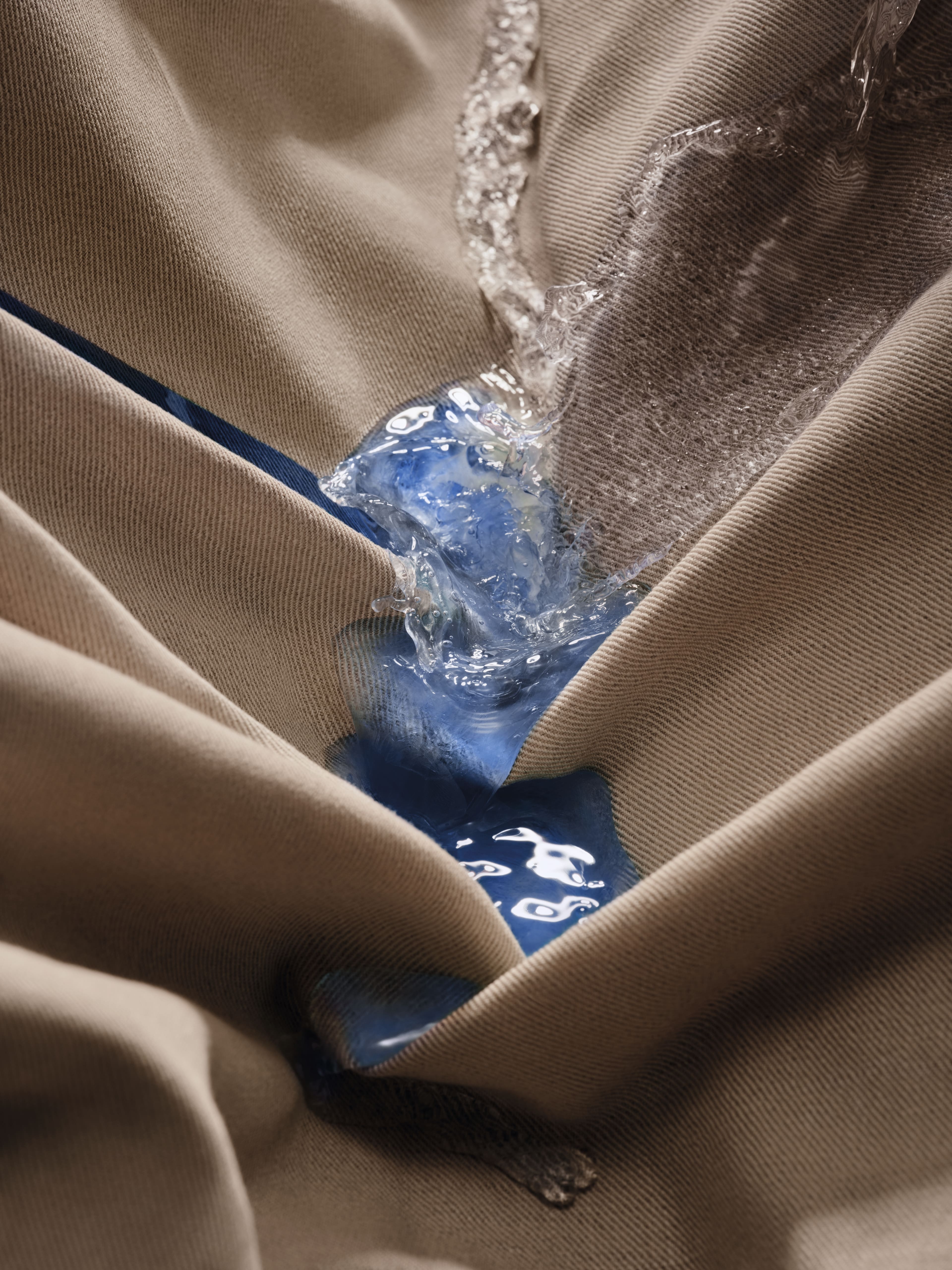 Gros plan sur un tissu beige où de l'eau claire et un liquide bleu éclaboussent les plis profonds, mettant en valeur les éclaboussures et la texture.