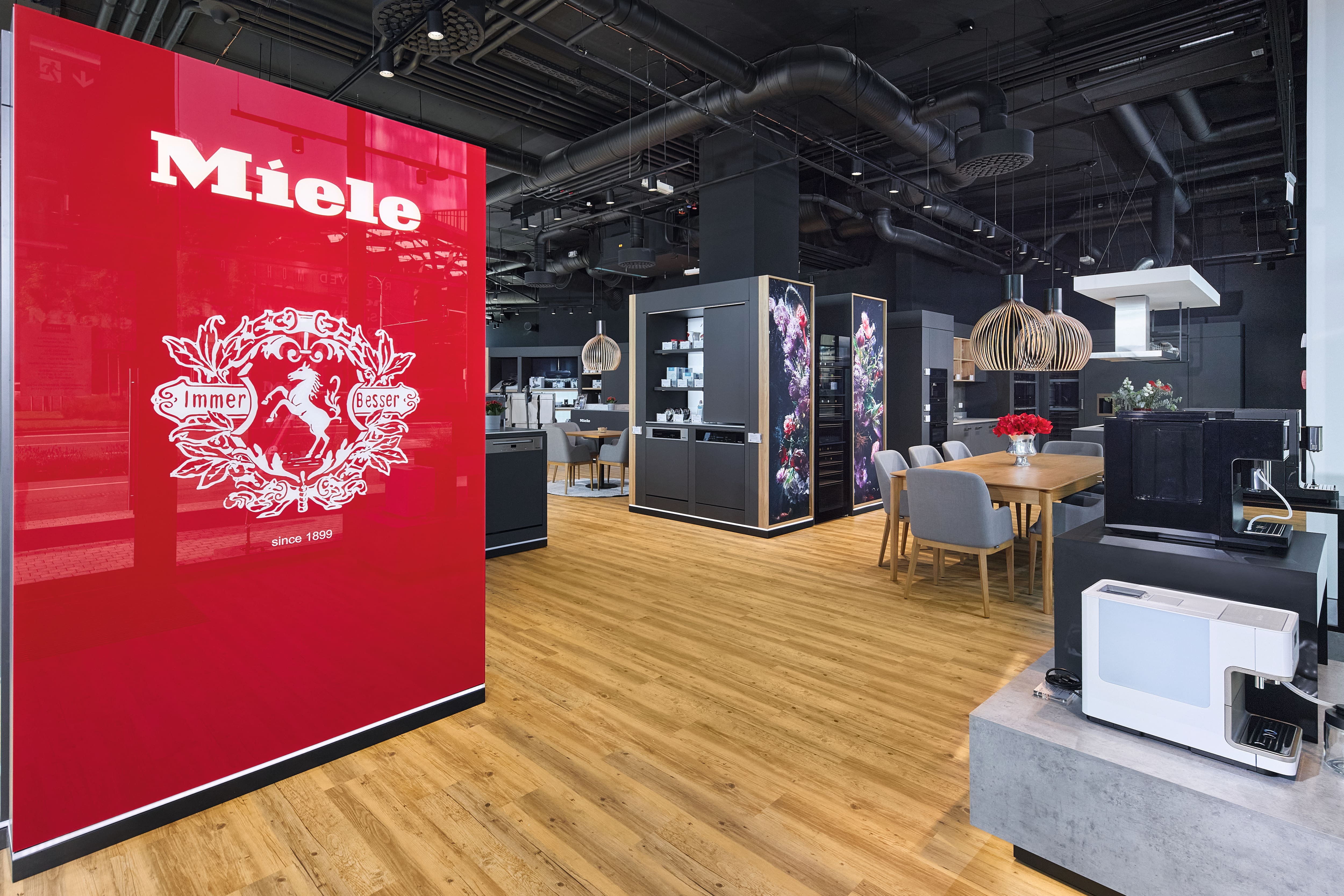 Moderný showroom Miele s kuchynskými spotrebičmi, demo stanicami a sedením, s výrazným červeným panelom značky.