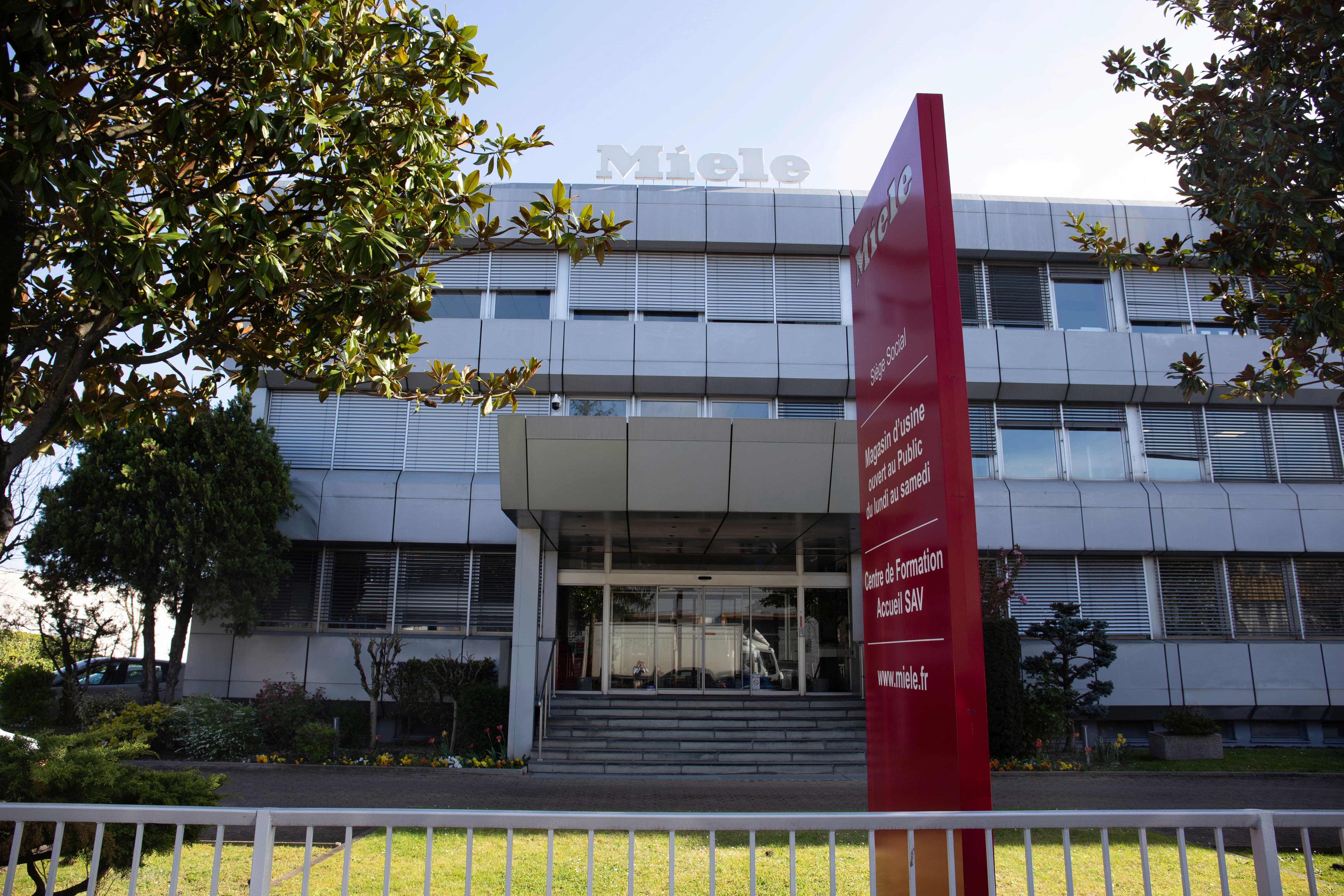 Vue de face d'un immeuble de bureaux Miele, avec une grande enseigne française rouge près de l'entrée, au milieu des arbres et de la verdure. Vue de face d'un immeuble de bureaux Miele, avec une grande enseigne française rouge près de l'entrée, au milieu des arbres et de la verdure.