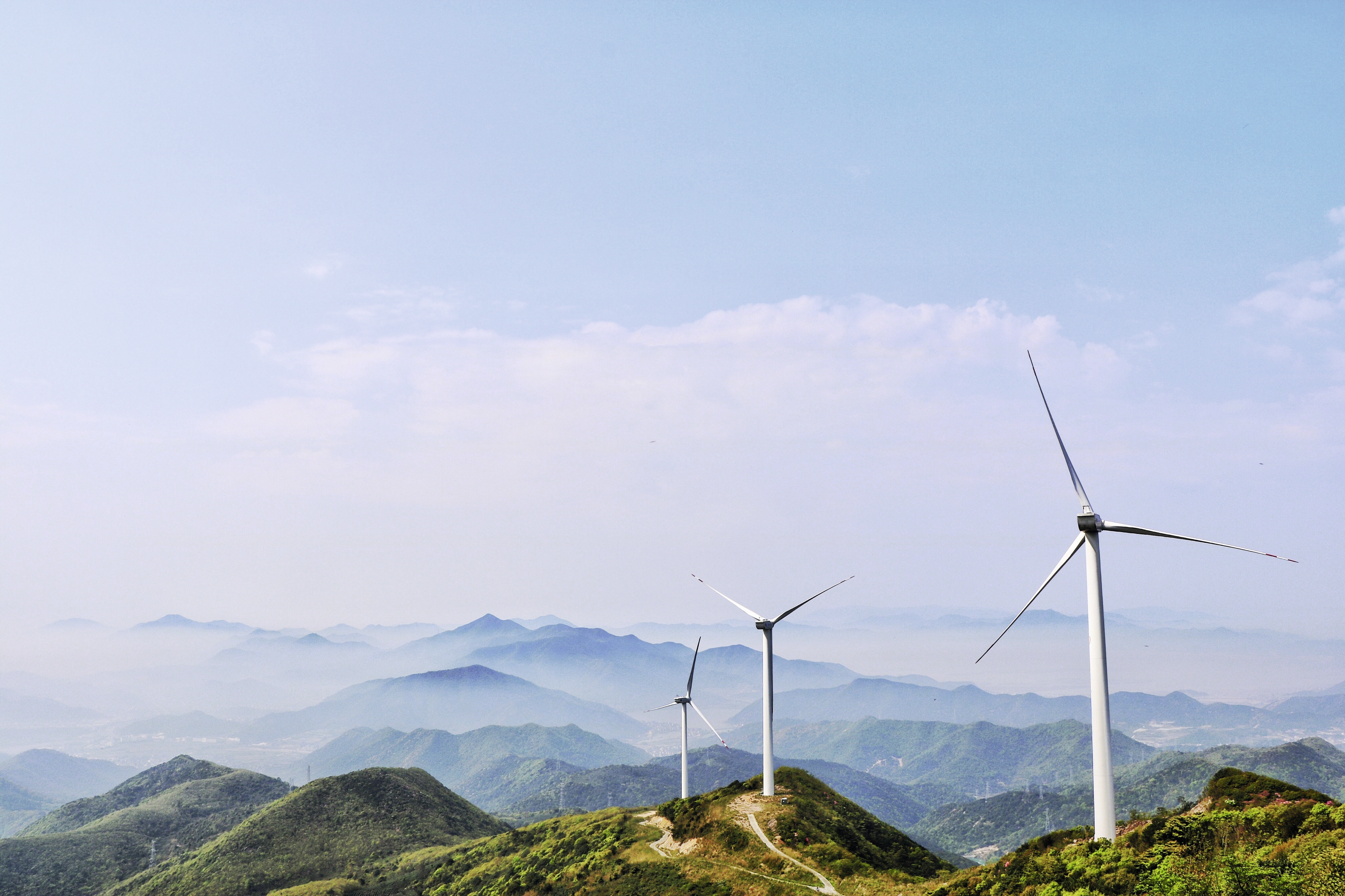 Wind turbines on green mountain ridges under a clear blue sky with distant hazy ranges.