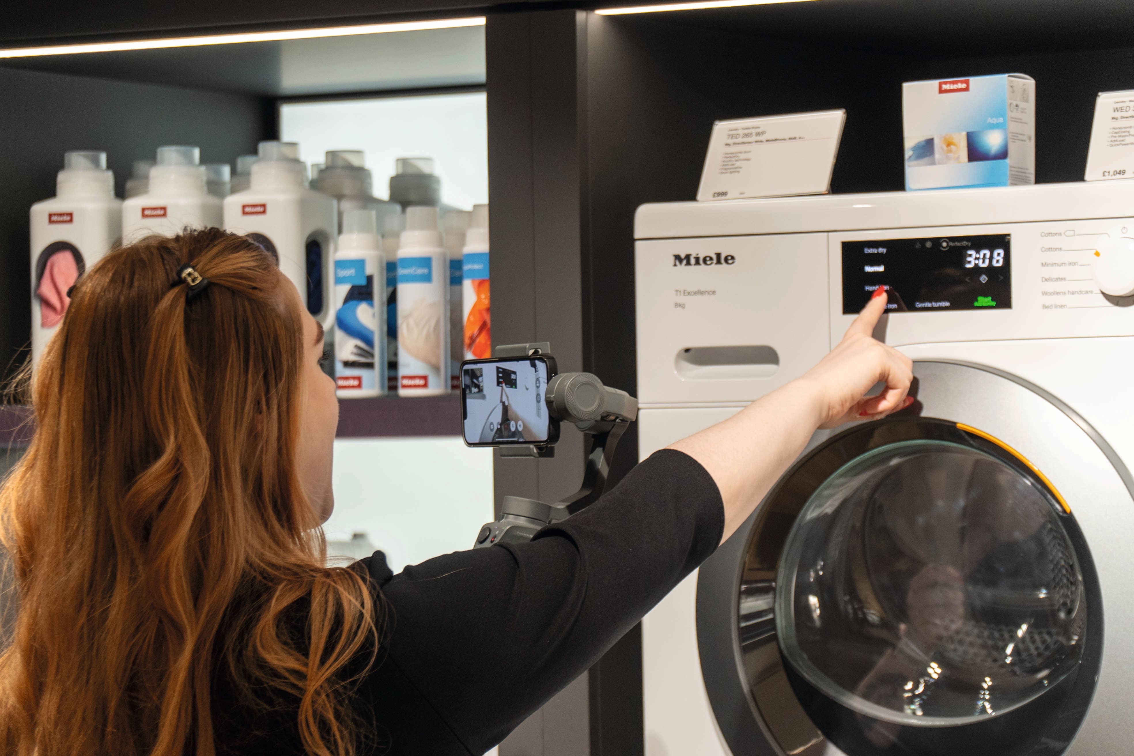 Clientul demonstrează o mașină de spălat Miele, selectând un ciclu în timp ce înregistrează cu un gimbal pentru smartphone într-un magazin. Clientul demonstrează o mașină de spălat Miele, selectând un ciclu în timp ce înregistrează cu un gimbal pentru smartphone într-un magazin.