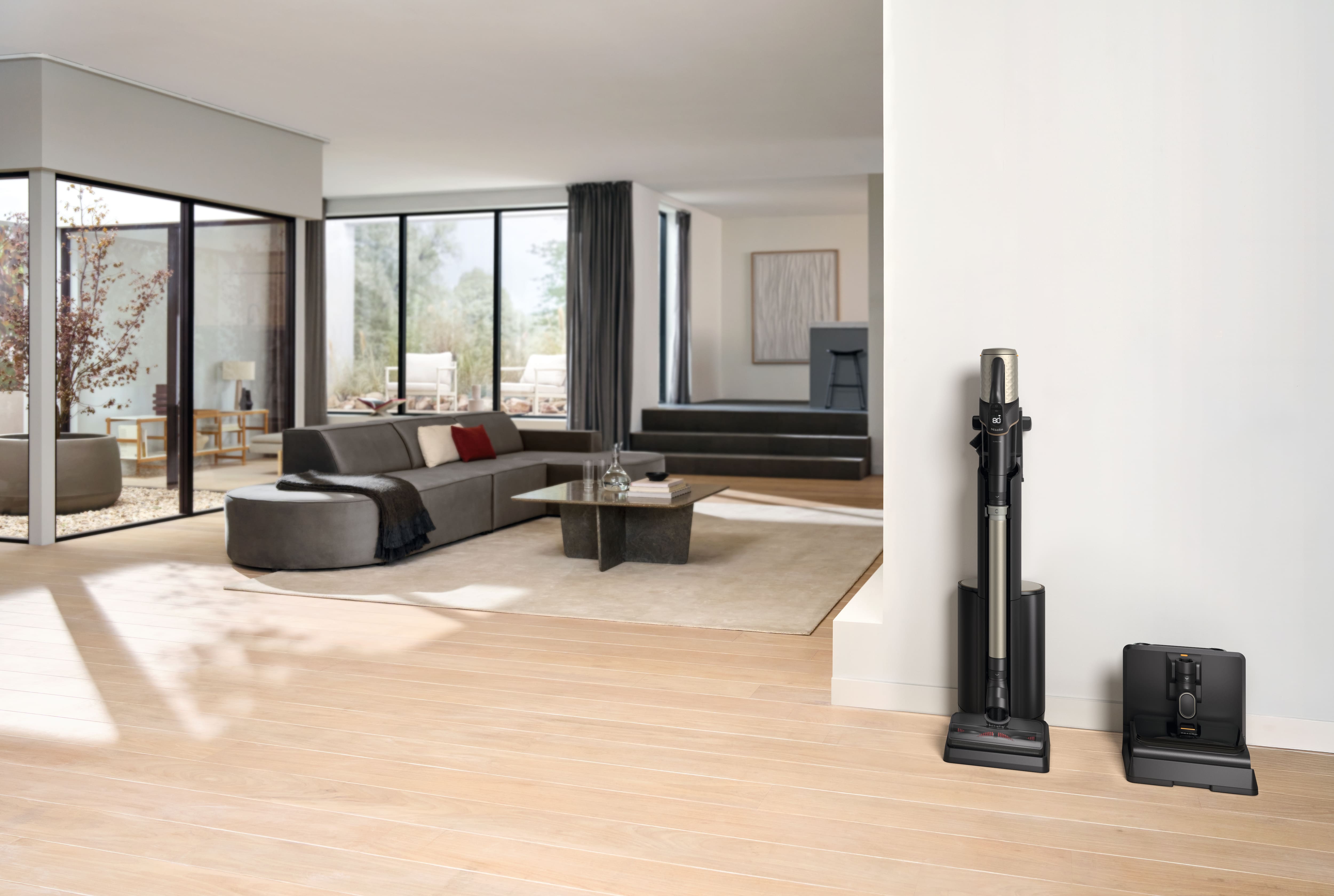 Helles, modernes Wohnzimmer mit grauem Ecksofa und Couchtisch; kabelloser Stabstaubsauger an der Wand angedockt. Helles, modernes Wohnzimmer mit grauem Ecksofa und Couchtisch; kabelloser Stabstaubsauger an der Wand angedockt.