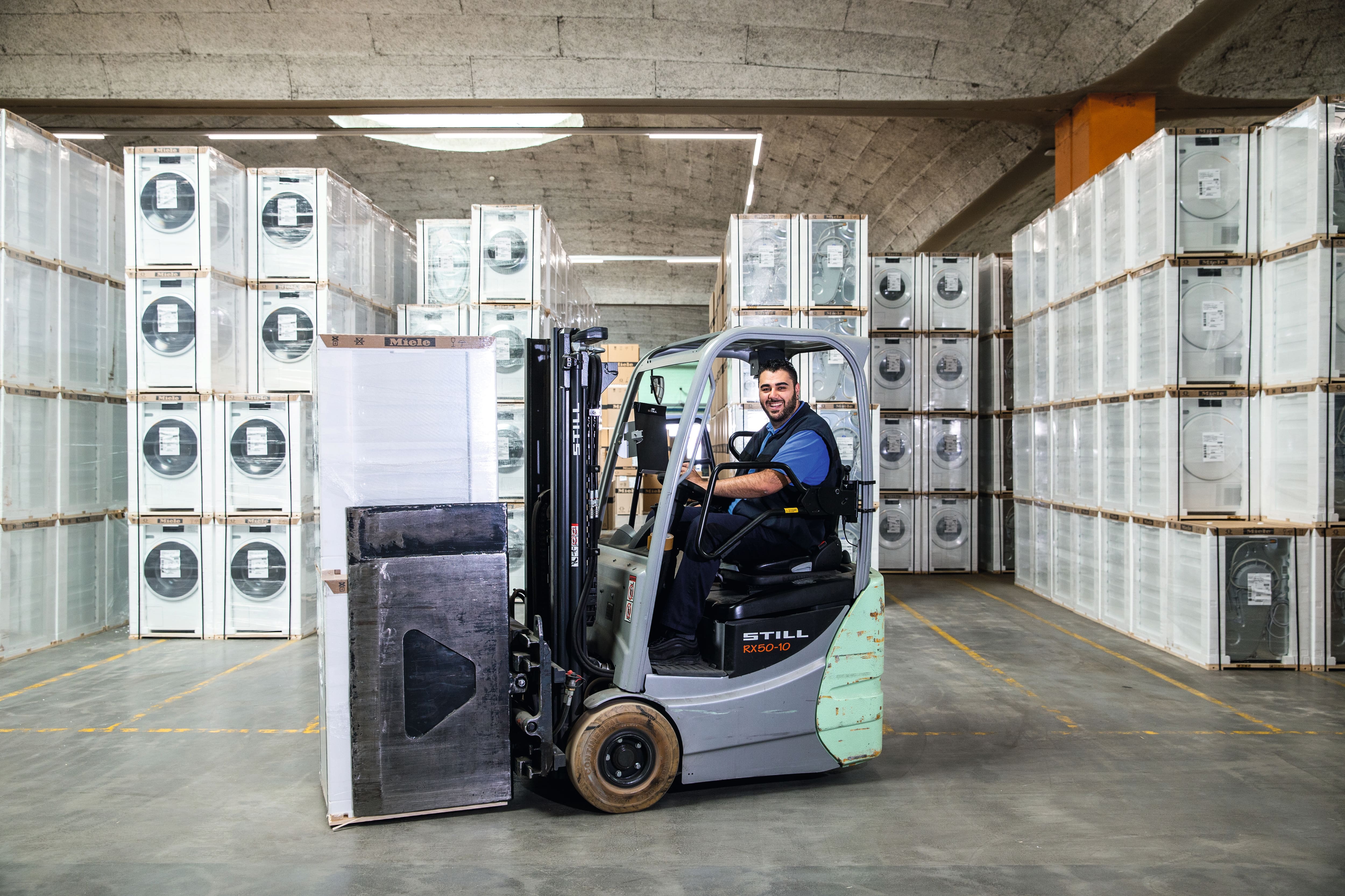 Lagerinnenraum mit einem Mitarbeiter, der einen Gabelstapler bedient und eine auf einer Palette verpackte Maschine transportiert, umgeben von hohen Stapeln verpackter Waschmaschinen.
