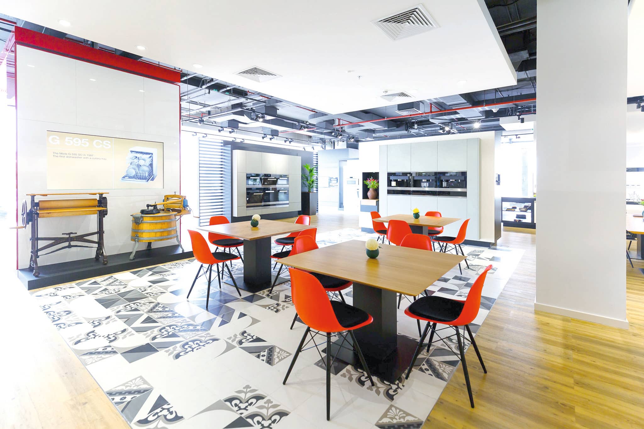 Bright modern showroom with wood tables, orange chairs, patterned tiles, and built-in kitchen appliances. Bright modern showroom with wood tables, orange chairs, patterned tiles, and built-in kitchen appliances.