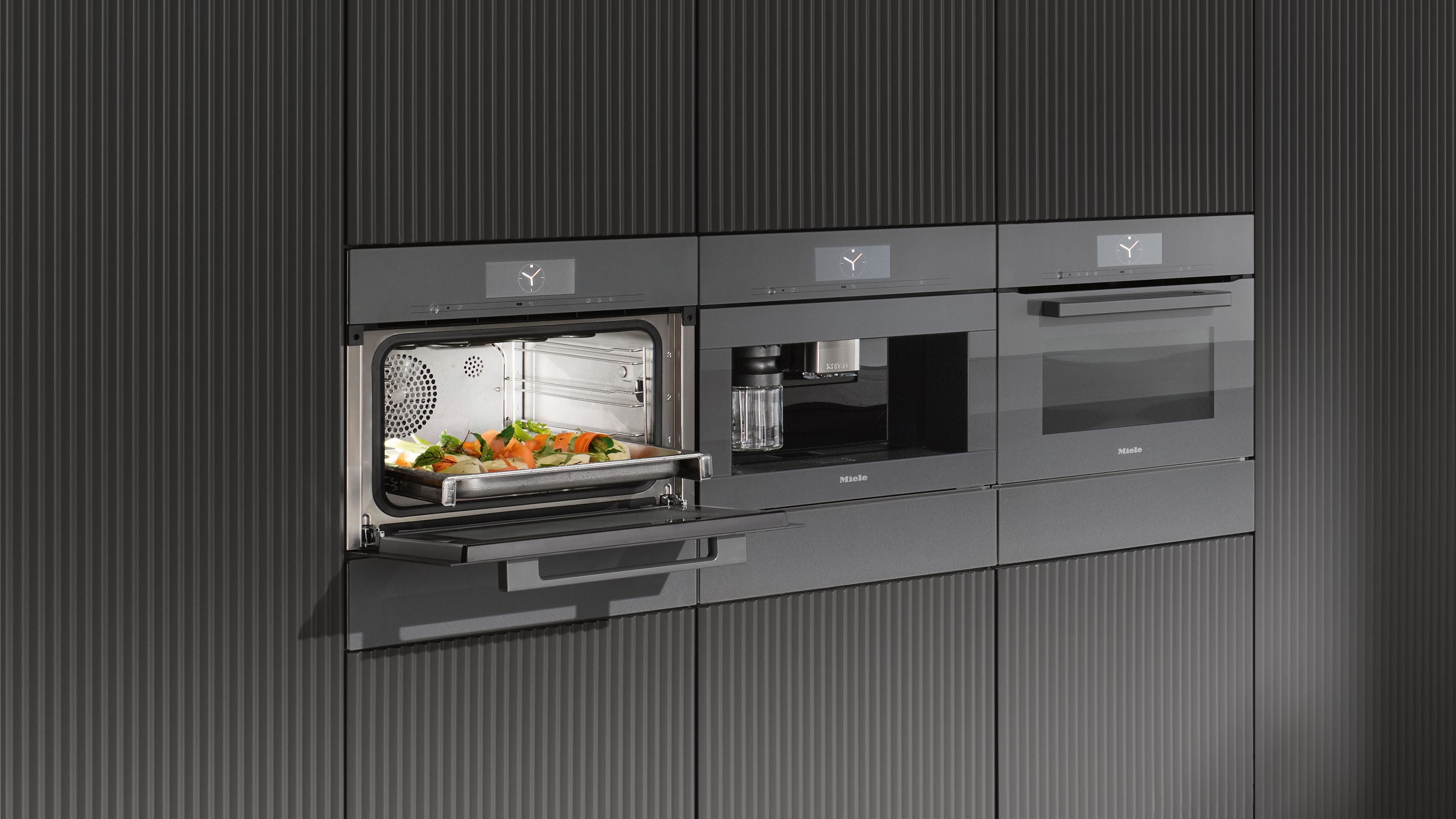 Sleek built‑in kitchen setup with three black appliances—an open steam oven holding a tray of mixed vegetables, a built‑in coffee machine, and an oven—set in ribbed dark cabinetry.
