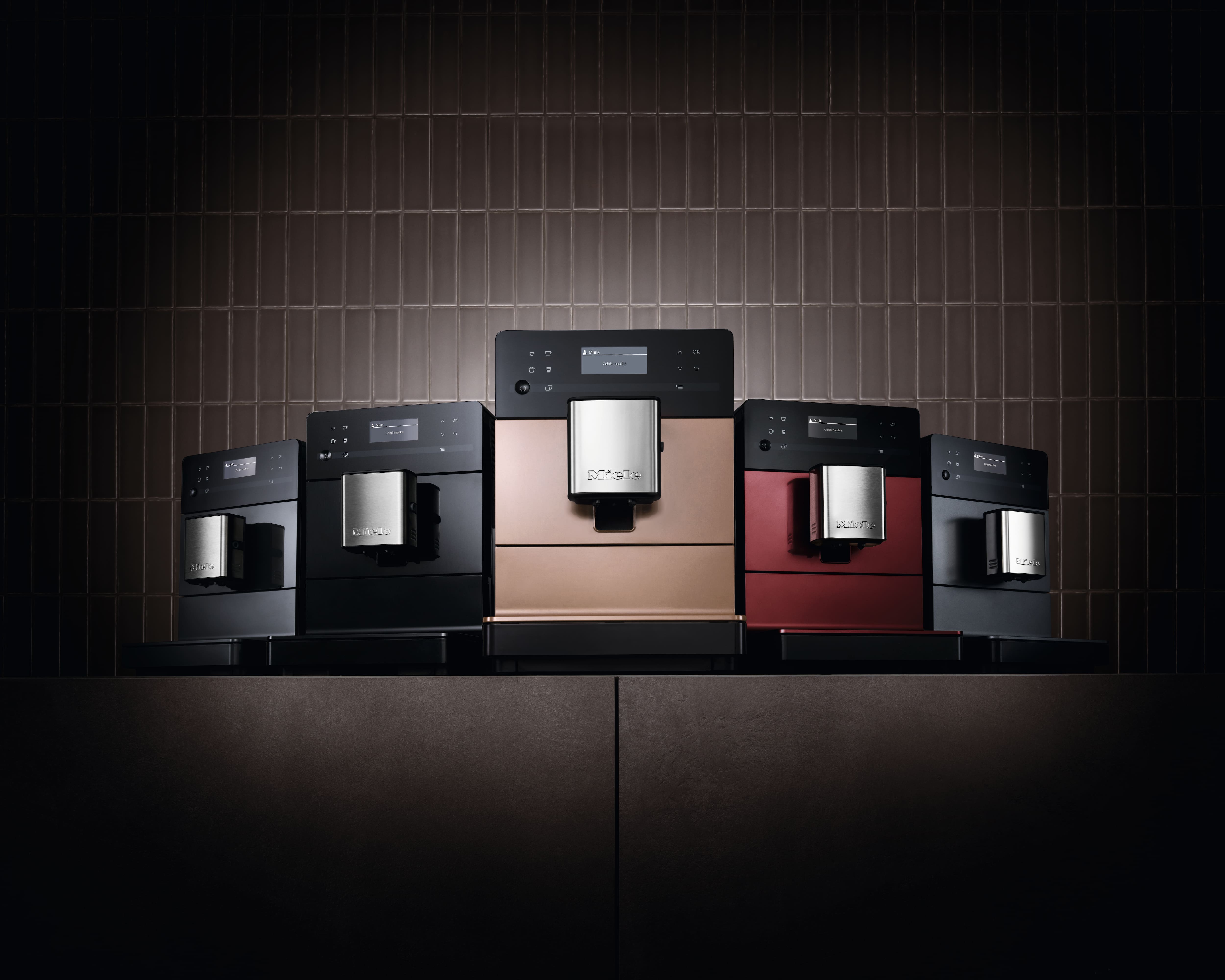 Gama de espressoare Miele pentru blat, în finisaje negru, bronz și roșu, pe un perete cu faianță închisă la culoare. Gama de espressoare Miele pentru blat, în finisaje negru, bronz și roșu, pe un perete cu faianță închisă la culoare.
