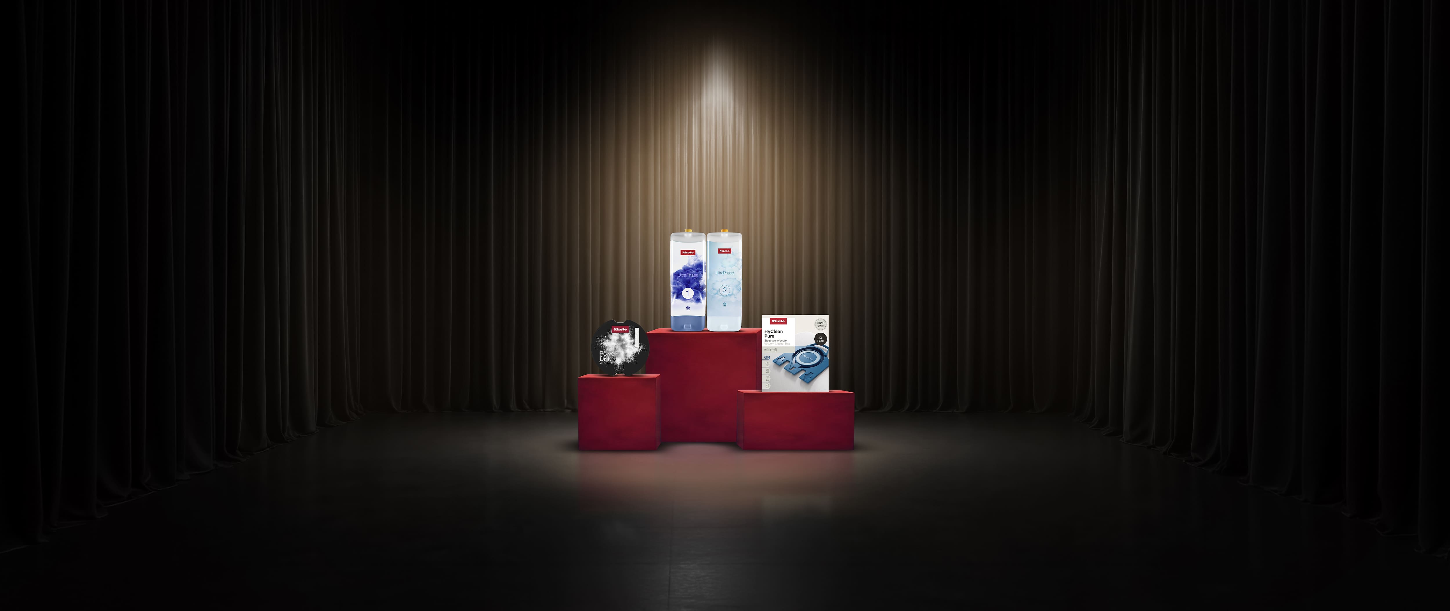 Une scène mise en valeur par des projecteurs, avec des podiums rouges présentant les consommables pour appareils électroménagers Miele, notamment des lessives et des sacs pour aspirateurs.