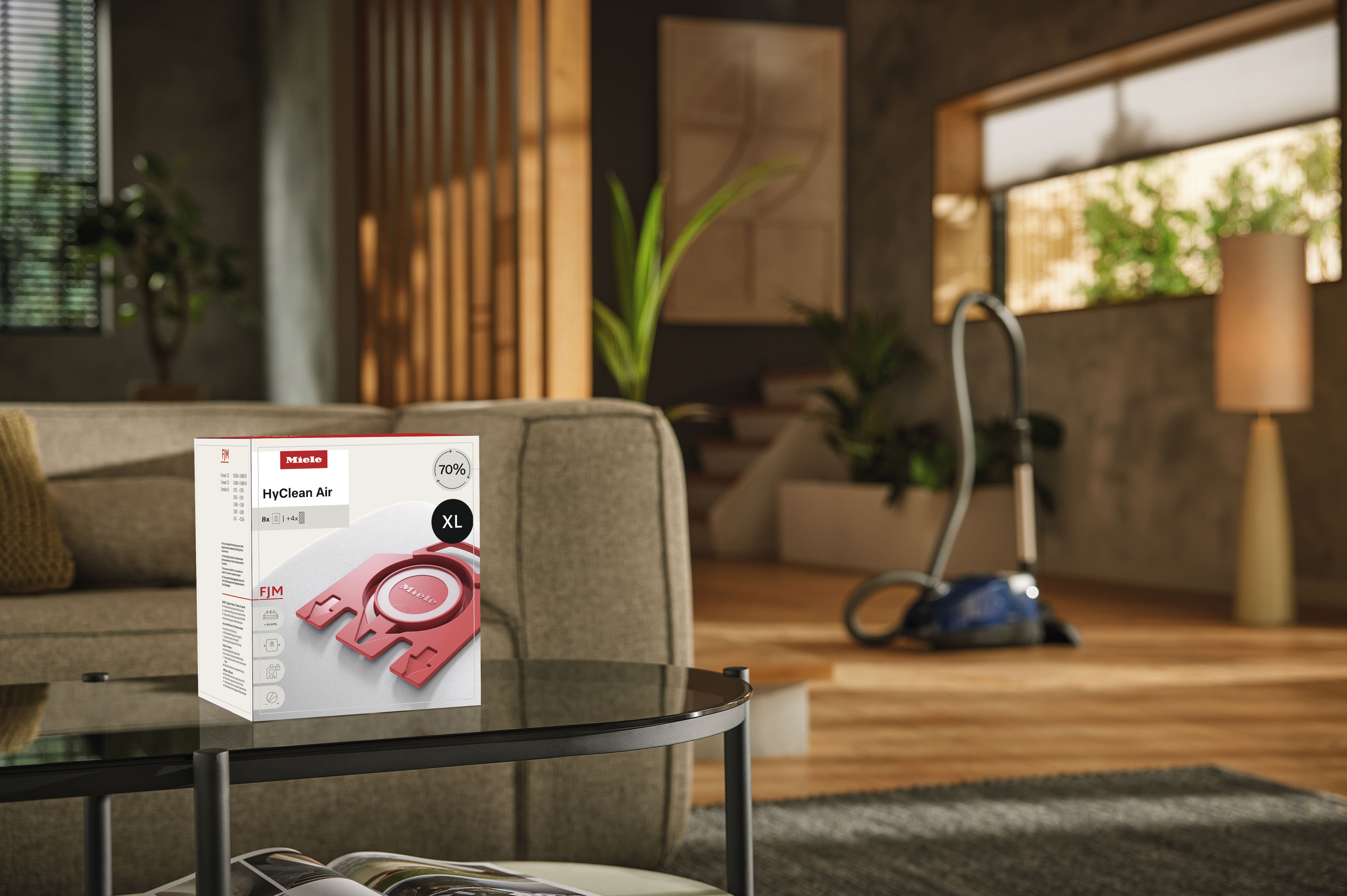 Modern living room with a box of Miele HyClean Air XL FJM vacuum bags on a glass coffee table, and a canister vacuum cleaner in the background near a large window. Modern living room with a box of Miele HyClean Air XL FJM vacuum bags on a glass coffee table, and a canister vacuum cleaner in the background near a large window.