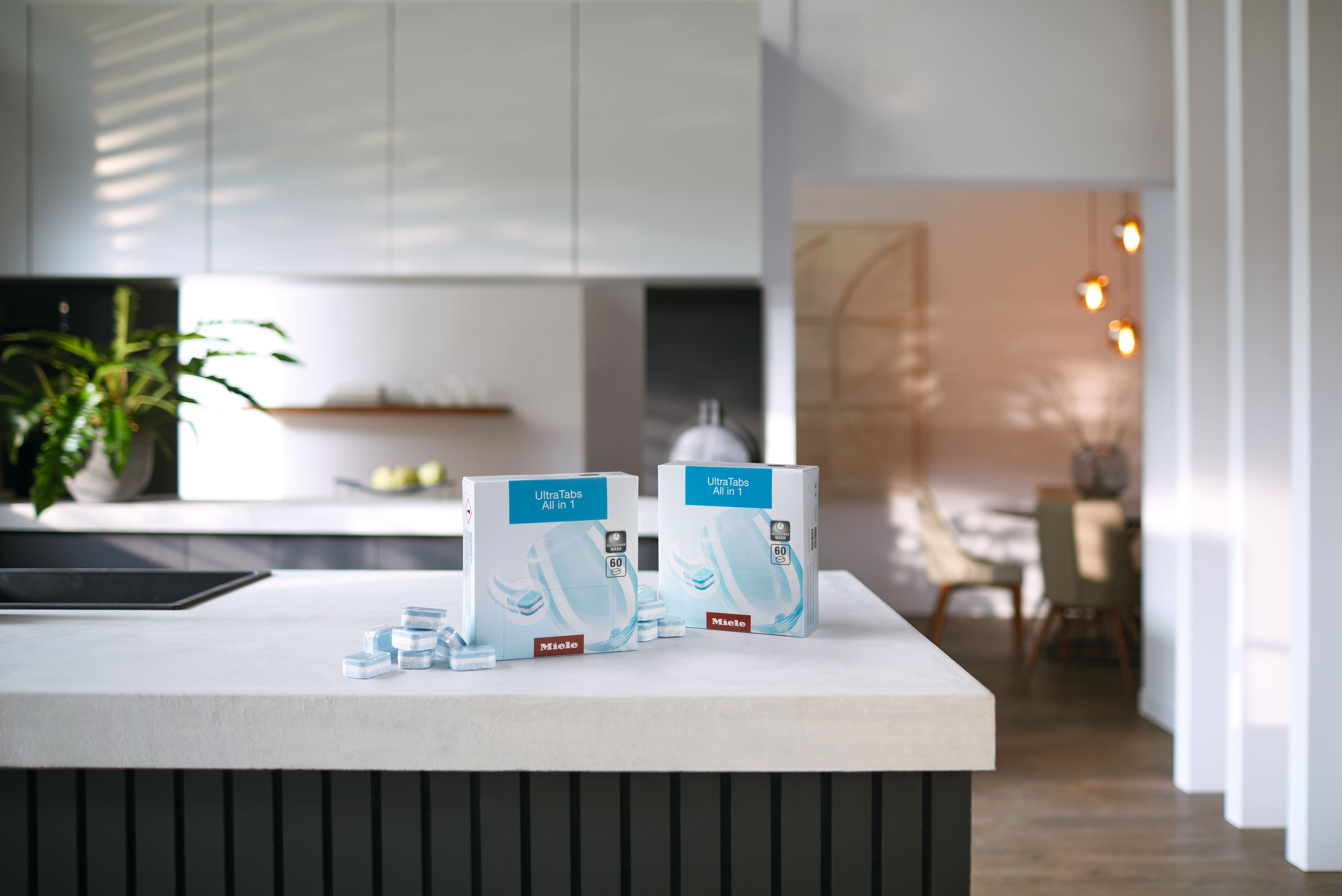 Due scatole di Miele UltraTabs All in 1 per lavastoviglie su un moderno piano cucina, con pastiglie blu sparse. Due scatole di Miele UltraTabs All in 1 per lavastoviglie su un moderno piano cucina, con pastiglie blu sparse.