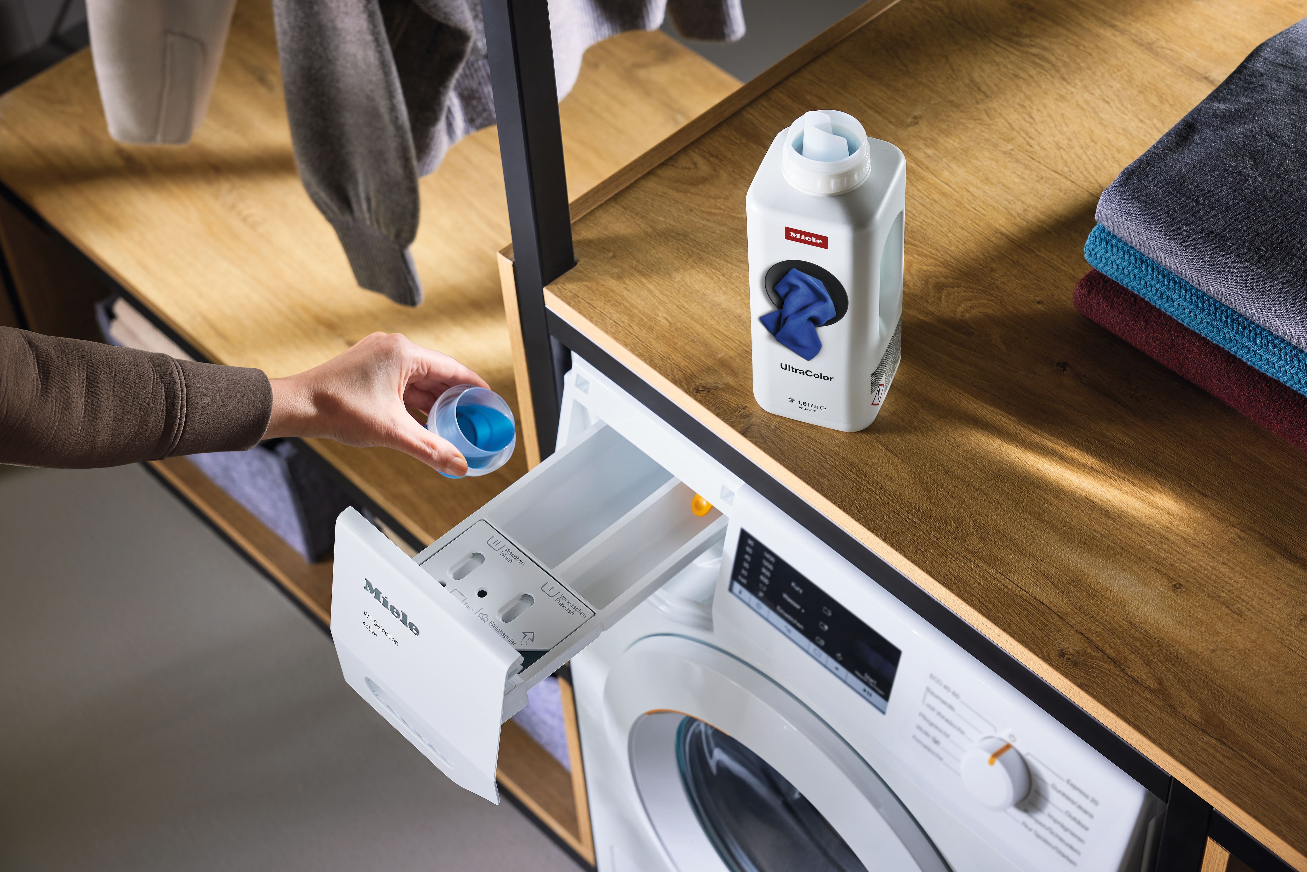 Persona versa del detersivo liquido blu con un tappo dosatore nel cassetto del detersivo di una lavatrice Miele. Accanto si trovano un flacone di detersivo Miele UltraColor e asciugamani piegati su un piano di lavoro in legno. Persona versa del detersivo liquido blu con un tappo dosatore nel cassetto del detersivo di una lavatrice Miele. Accanto si trovano un flacone di detersivo Miele UltraColor e asciugamani piegati su un piano di lavoro in legno.