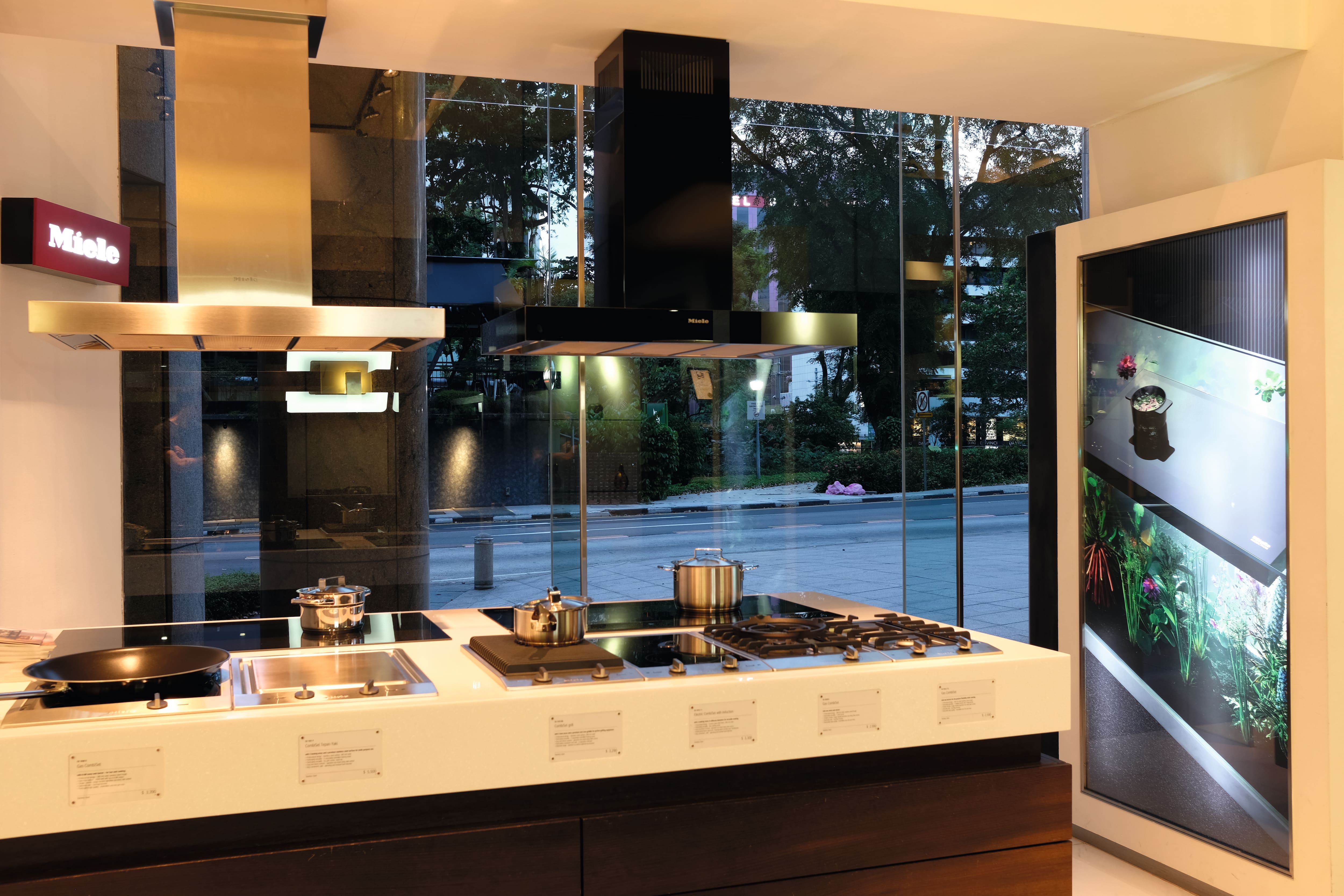 Modern kitchen showroom displays induction and gas cooktops with stainless range hoods, pots, and street view outside.