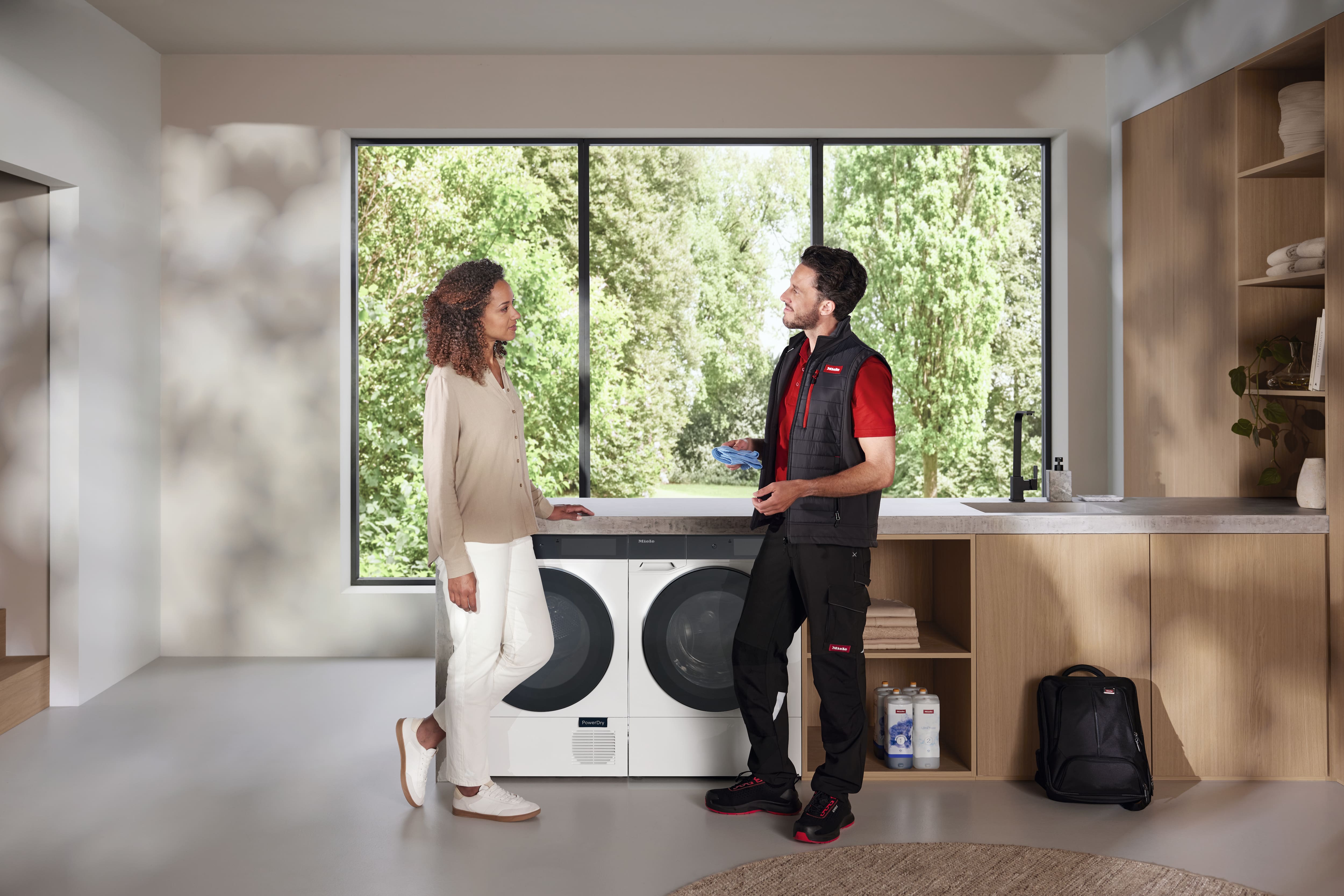 Appliance technician advising homeowner beside front-load washer and dryer in a bright, modern laundry room.