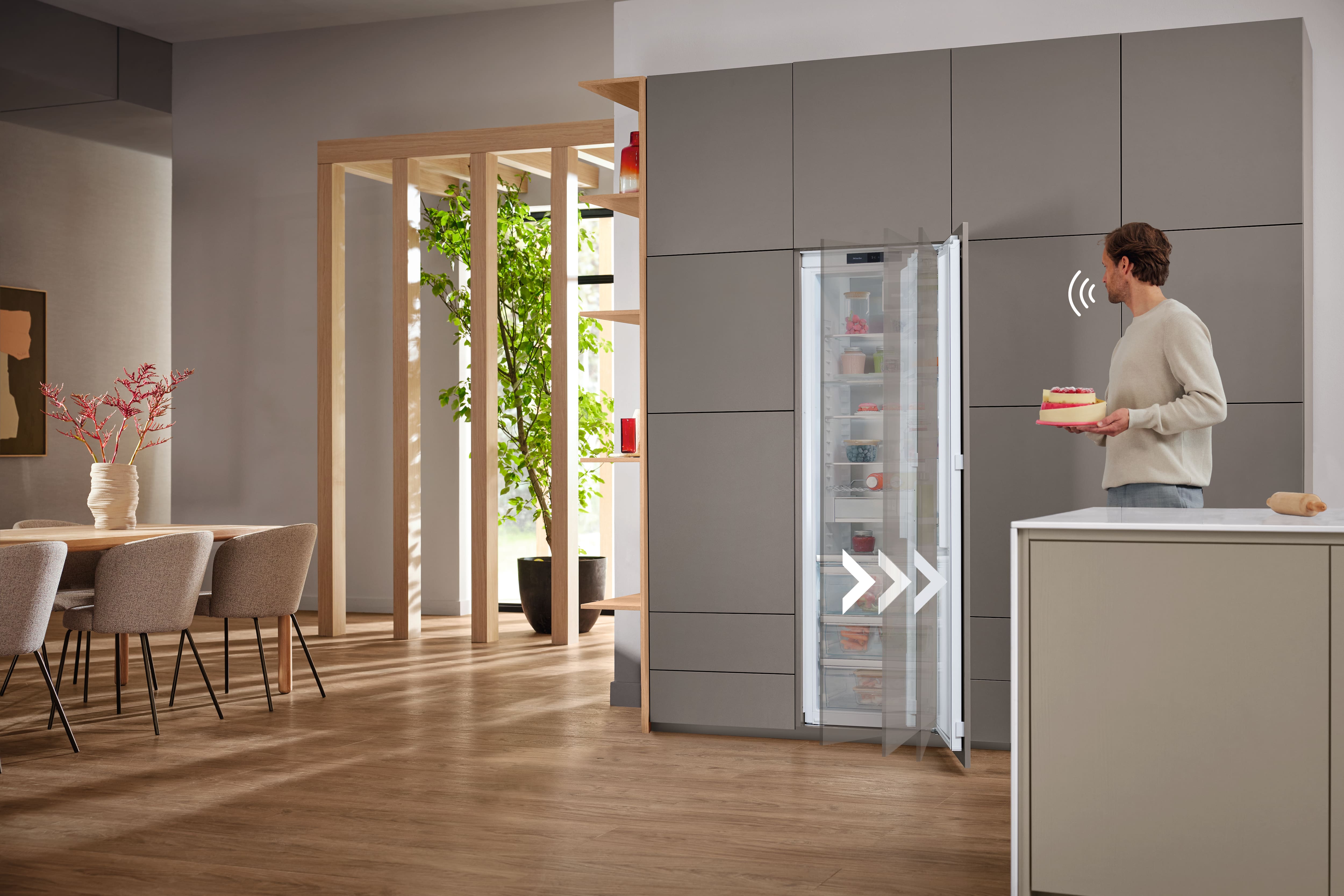 Modern kitchen with built-in smart fridge opening via gesture as a man holding a cake stands nearby. Modern kitchen with built-in smart fridge opening via gesture as a man holding a cake stands nearby.