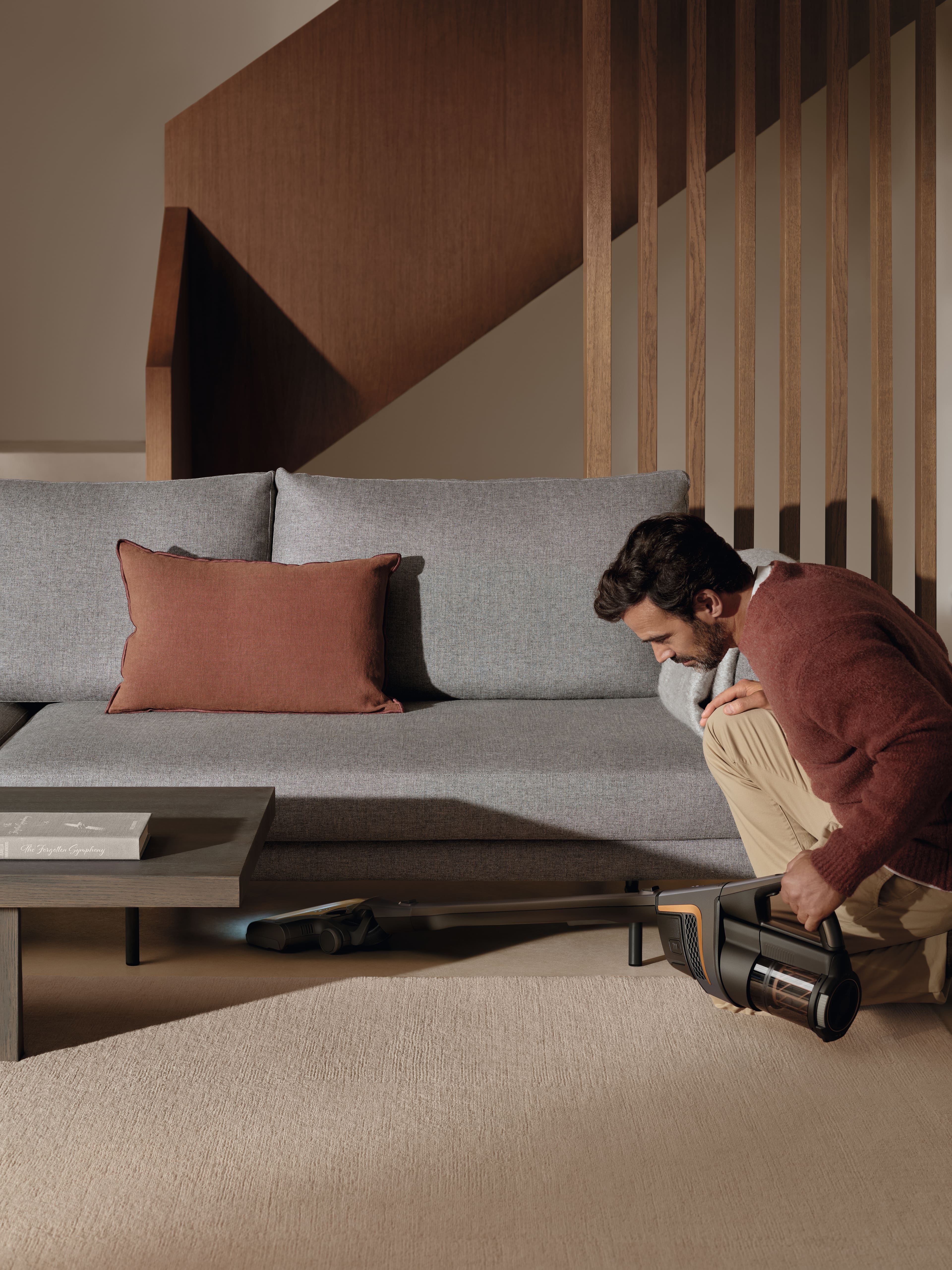Person using a Miele cordless stick vacuum to reach under a low coffee table in a modern living room with sofa. Person using a Miele cordless stick vacuum to reach under a low coffee table in a modern living room with sofa.