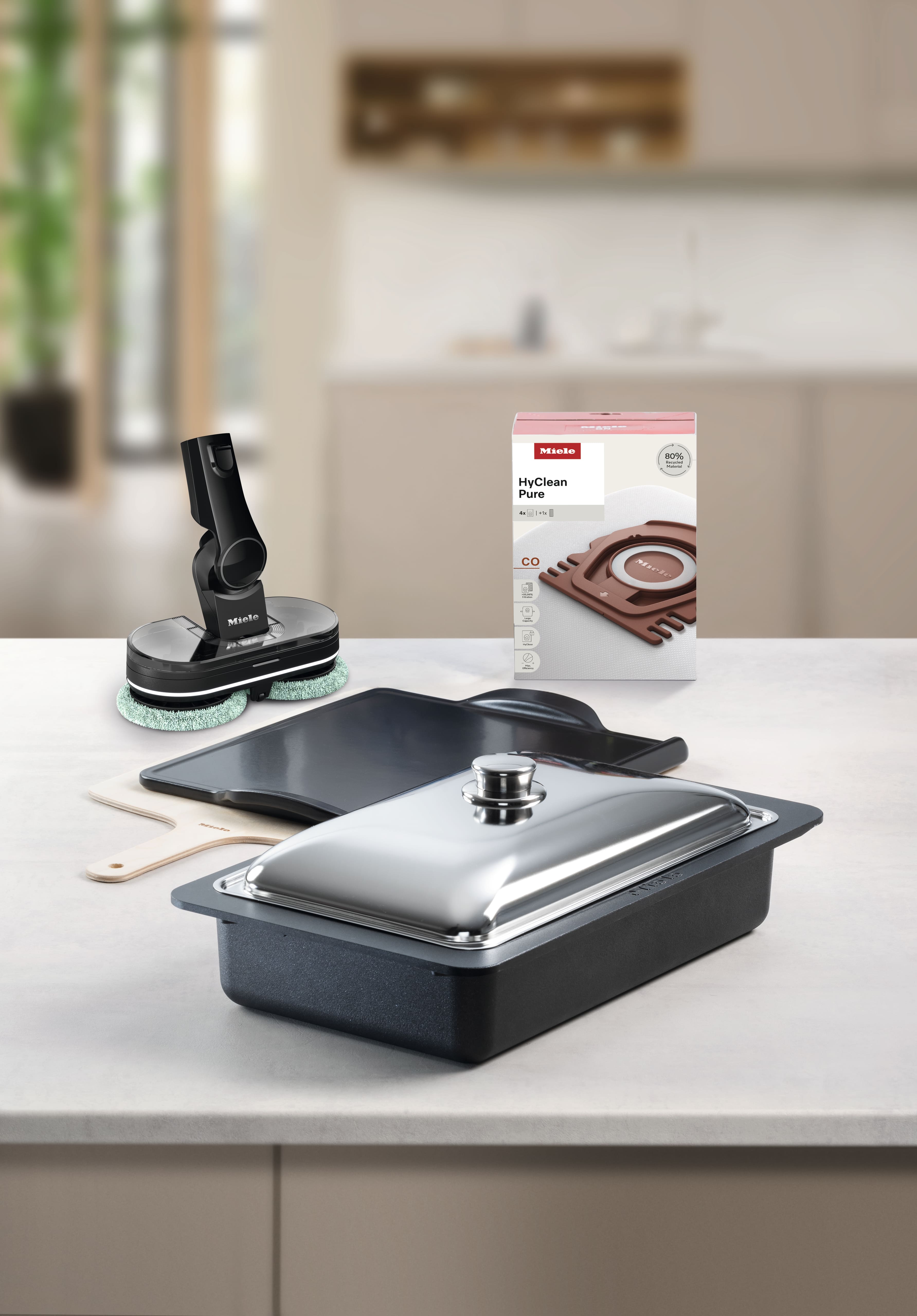 Assortiment d'accessoires Miele sur un plan de travail de cuisine : plat à rôtir avec couvercle en acier, plaques de four, tête de brosse et sacs d'aspirateur HyClean Pure. Assortiment d'accessoires Miele sur un plan de travail de cuisine : plat à rôtir avec couvercle en acier, plaques de four, tête de brosse et sacs d'aspirateur HyClean Pure.
