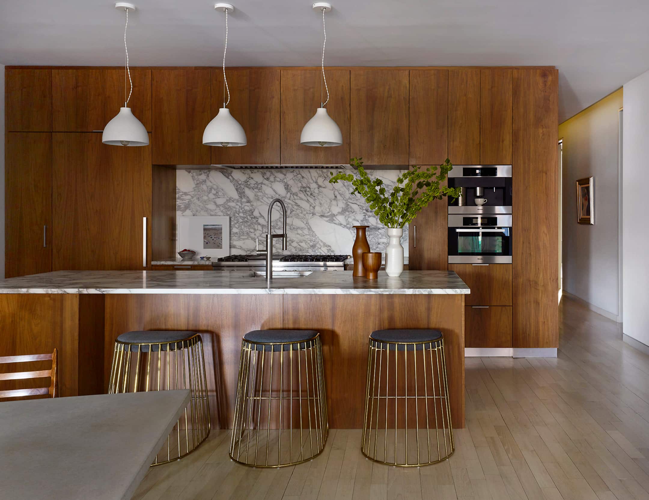 Modern kitchen with walnut cabinets, marble island and backsplash, brass wire stools, and white pendant lights. Modern kitchen with walnut cabinets, marble island and backsplash, brass wire stools, and white pendant lights.
