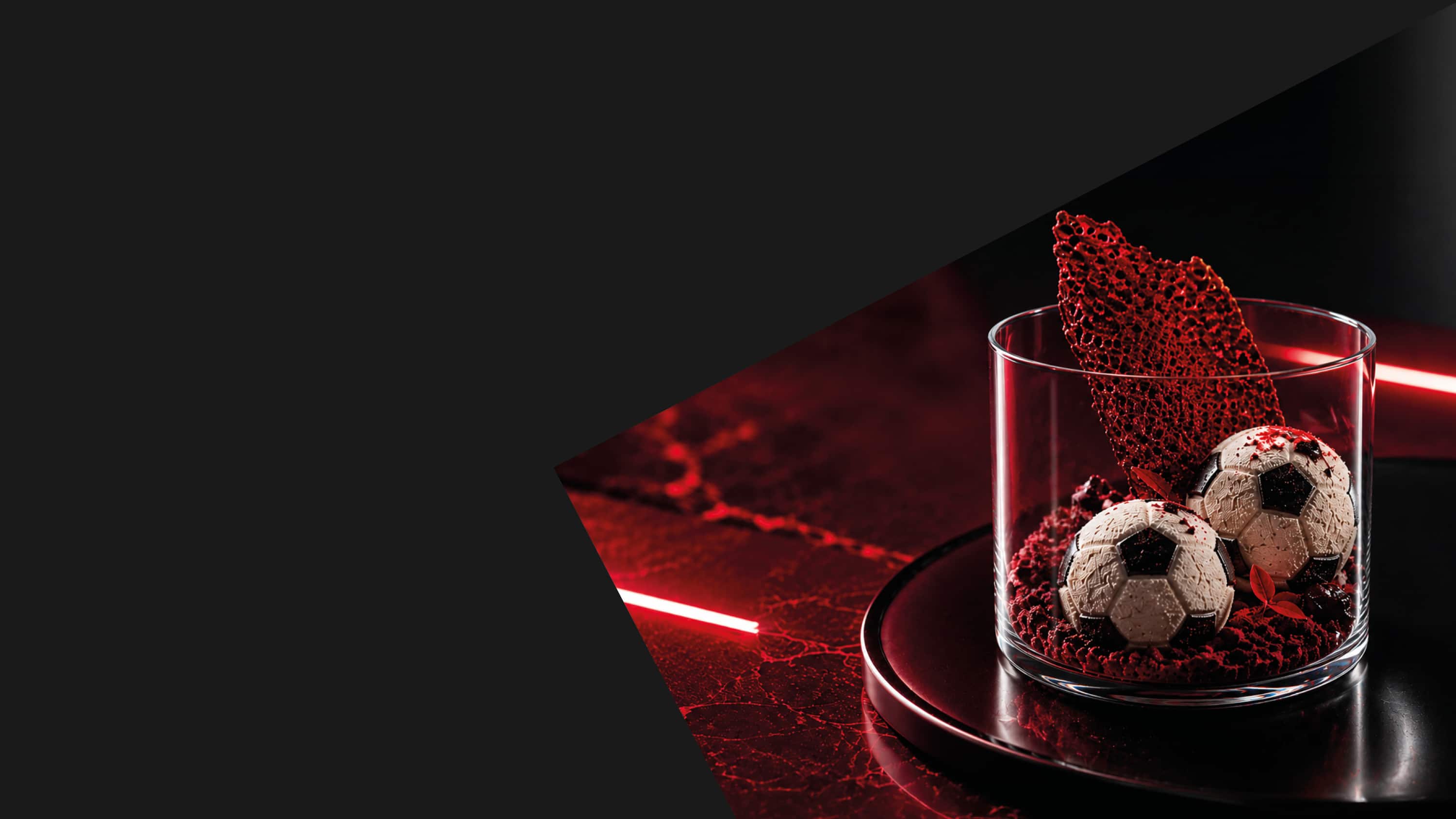 Glasdessert mit zwei schokoladenen Pralinen in Fussball-Optik auf rotem Crumble. Dramatische rote Neonbeleuchtung und schwarzer Untergrund sorgen für eine moderne, kontrastreiche Inszenierung. Glasdessert mit zwei schokoladenen Pralinen in Fussball-Optik auf rotem Crumble. Dramatische rote Neonbeleuchtung und schwarzer Untergrund sorgen für eine moderne, kontrastreiche Inszenierung.